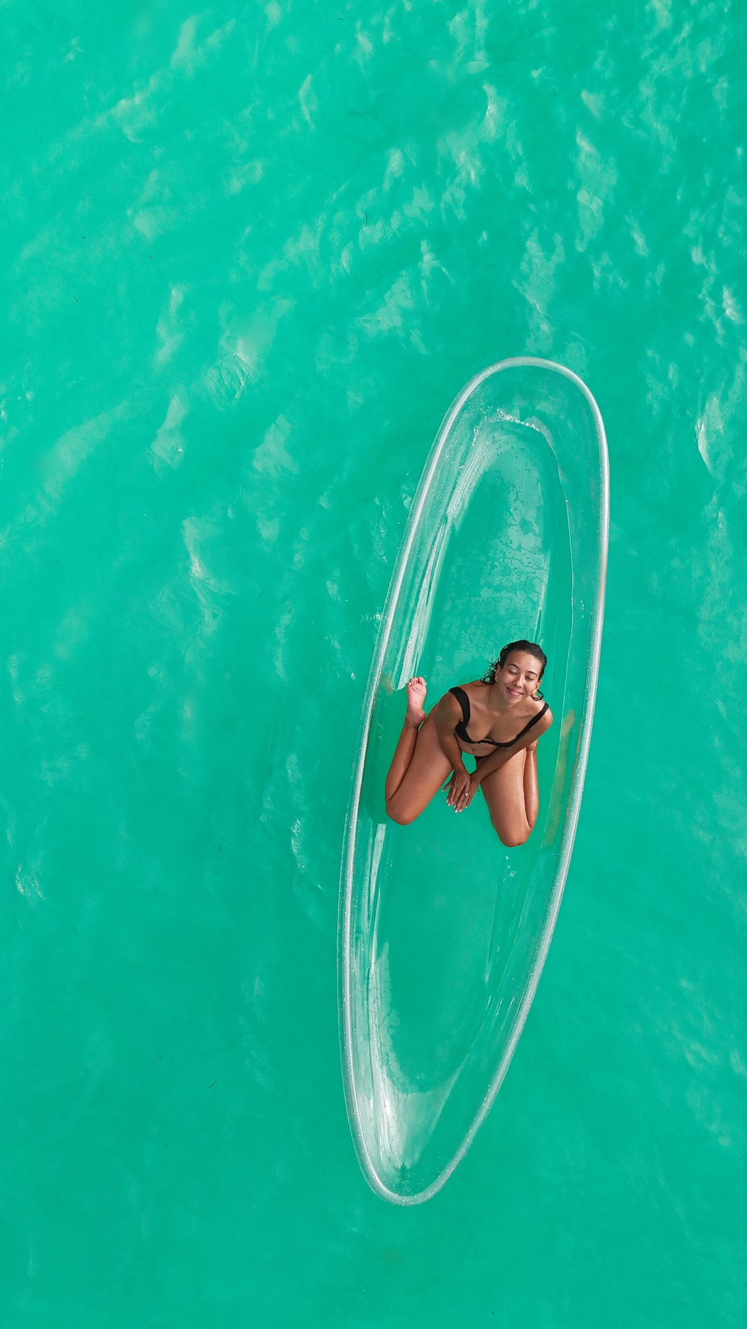 Aerial view of a transparent kayak floating on bright turquoise tropical waters with a person relaxing.