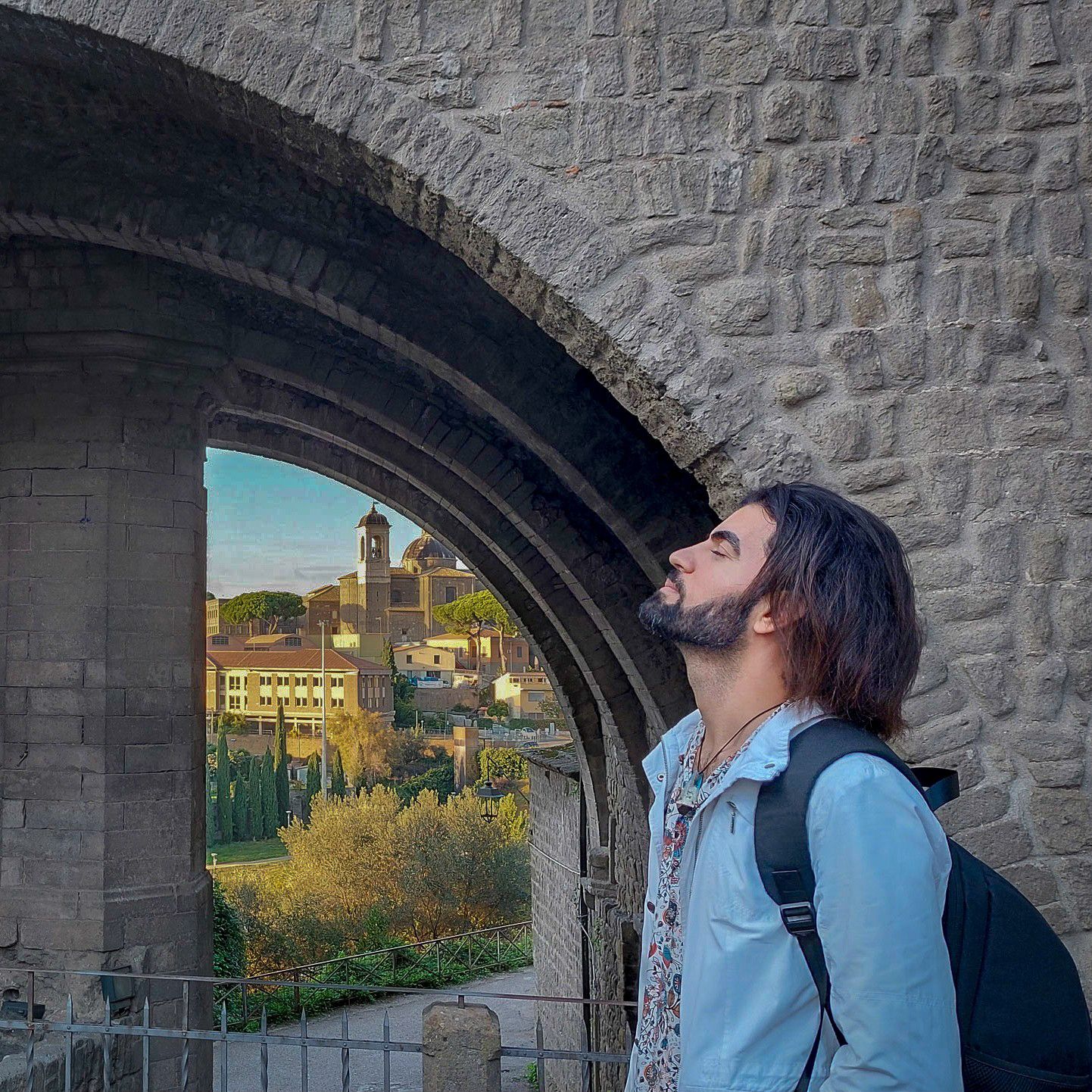 Man with long hair and backpack gazes up through a stone arch with scenic European cityscape in background.