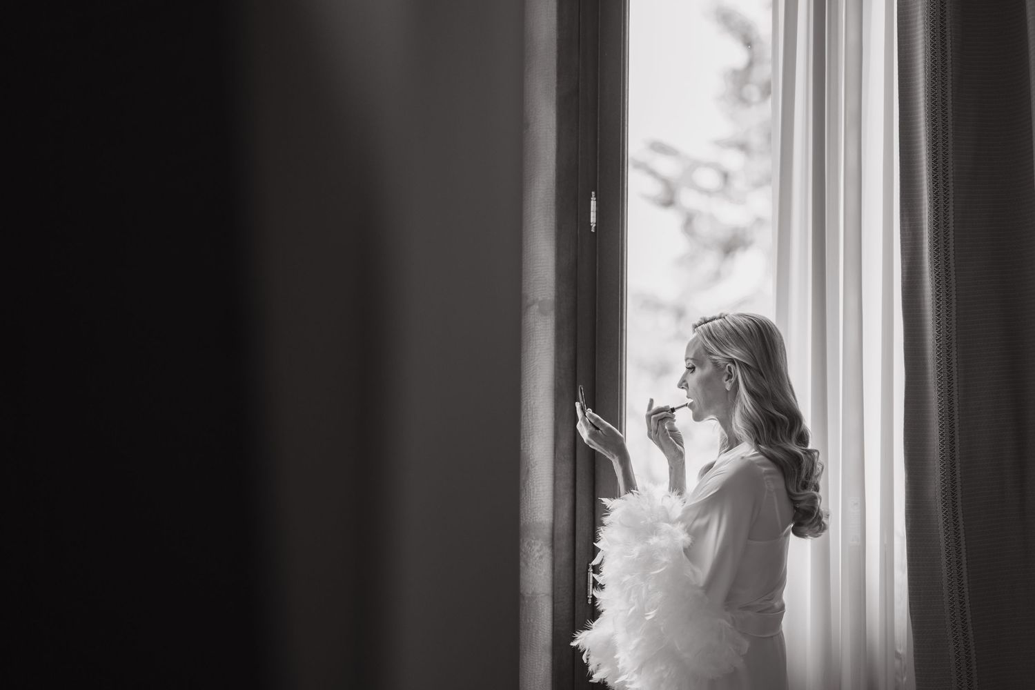 Black and white photo of bride applying makeup by a window in a feathered robe before her wedding.