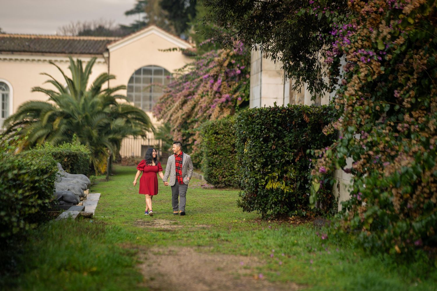 Couple walking hand in hand through a lush garden path beside a white villa with blooming purple flowers.