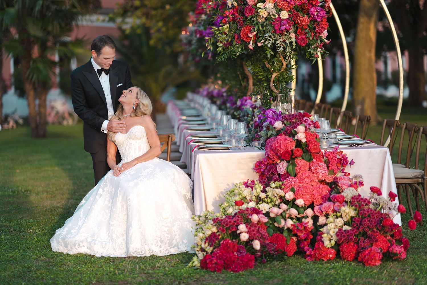 Bride in ball gown seated beside groom at outdoor wedding reception with vibrant floral arrangements.