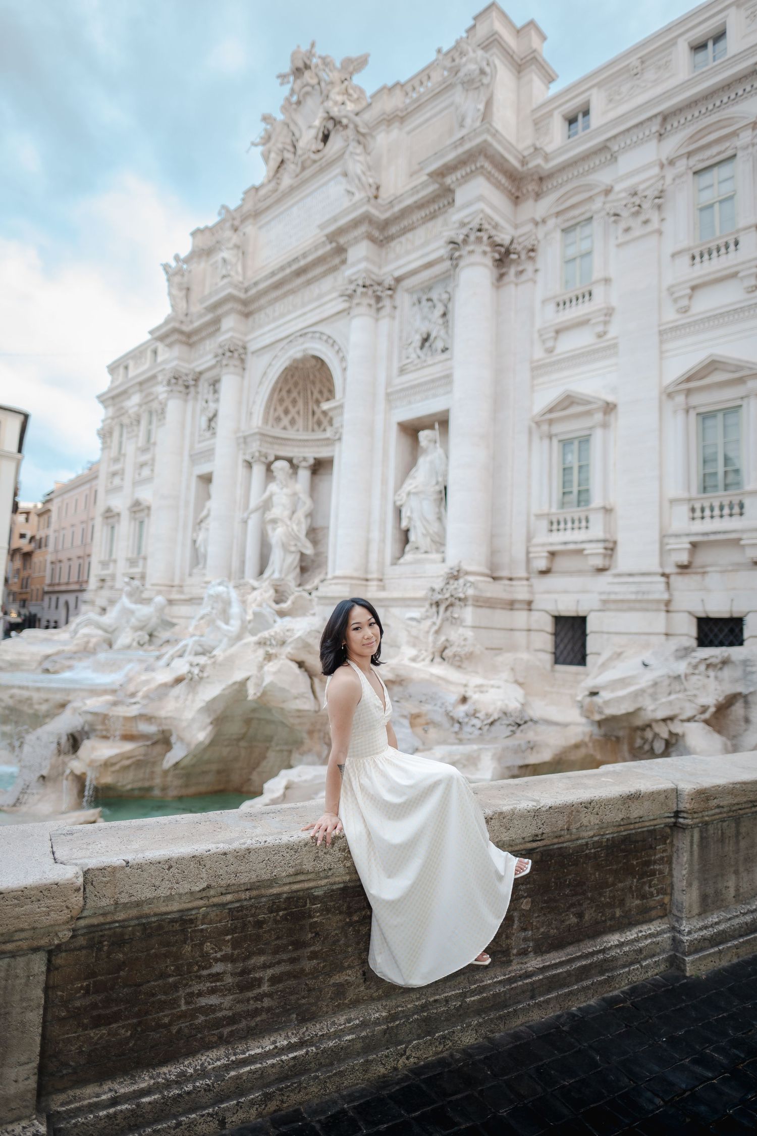 Woman in white dress sitting at Rome's Trevi Fountain, posing elegantly against the baroque marble facade.