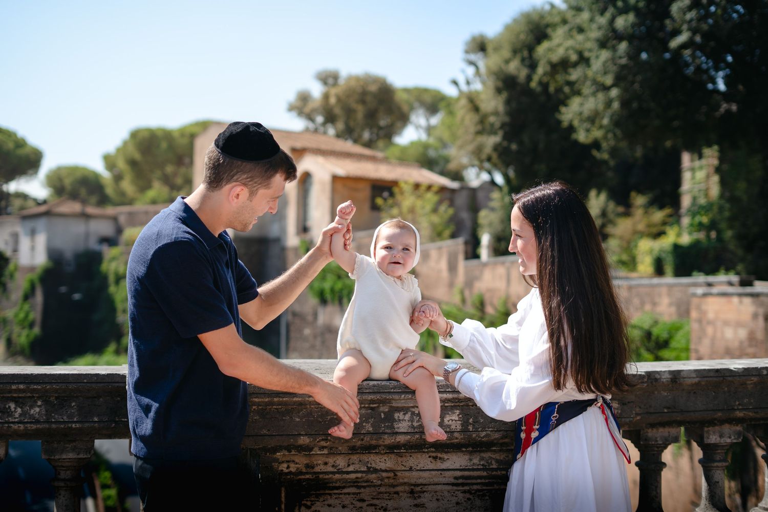 Young family with baby sitting on stone wall in Rome, Italy, with lush greenery and historic ruins in background.
