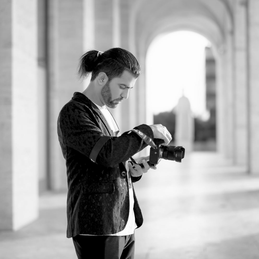 Black and white photo of a stylish man with a bun hairstyle checking his camera between elegant columns.