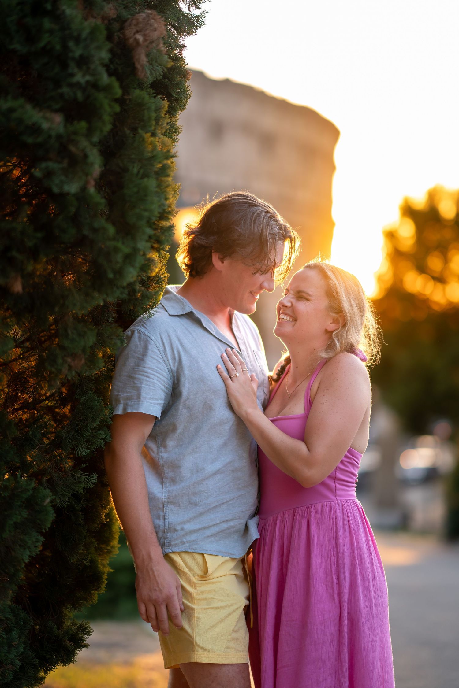 Couple sharing a romantic moment at golden hour near green trees, woman in pink dress smiling at man in blue shirt.