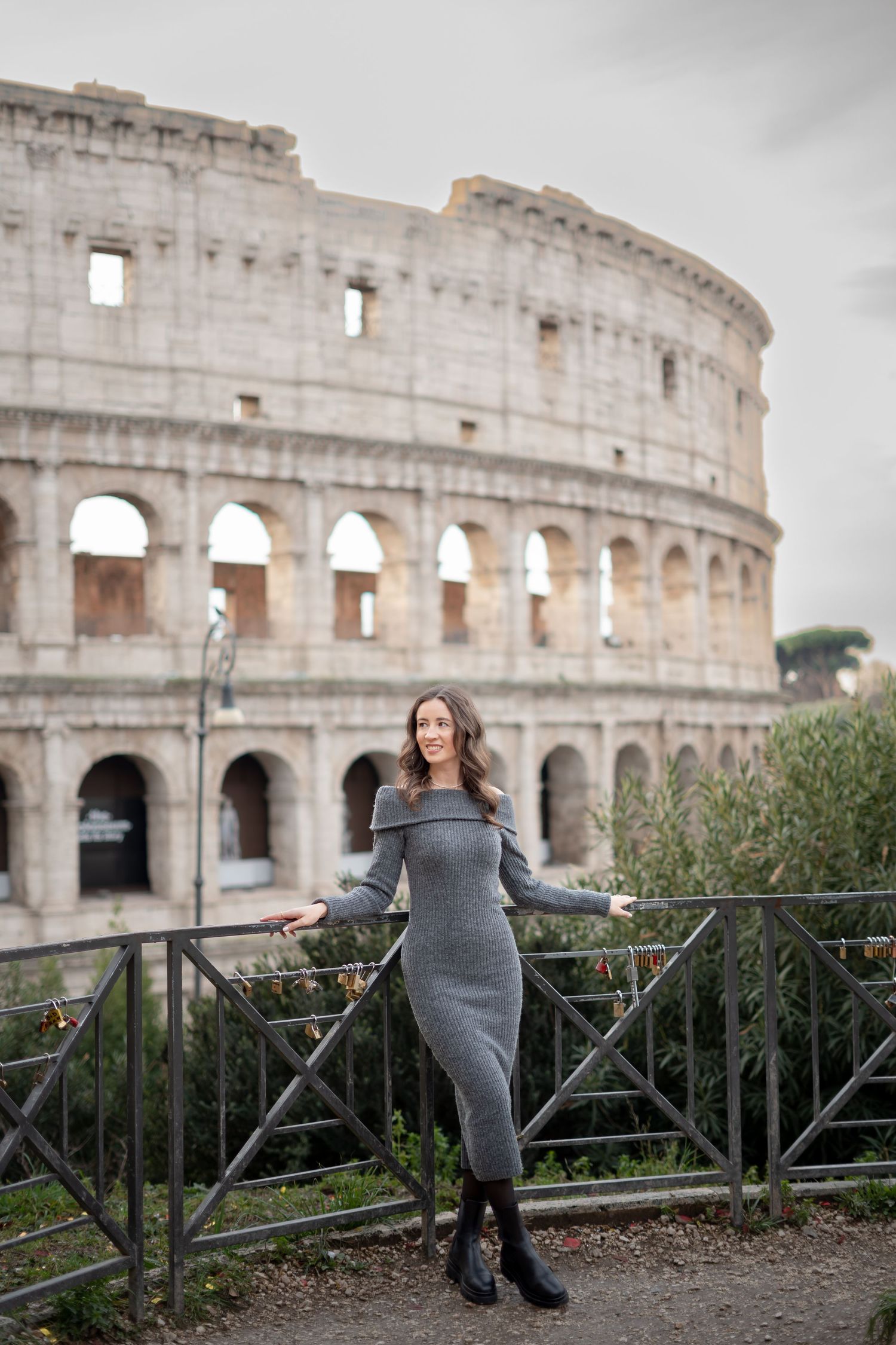 Woman in gray off-shoulder dress posing at metal fence with Rome's iconic Colosseum in background.