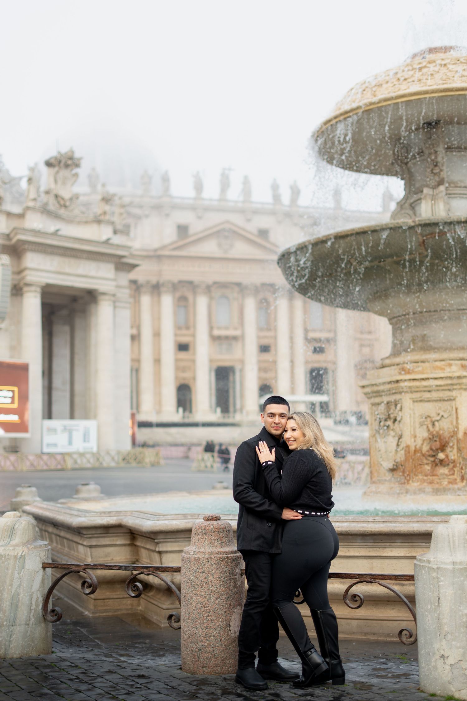 Couple in black outfits embracing by a fountain in St. Peter's Square, Vatican City, on a misty day.