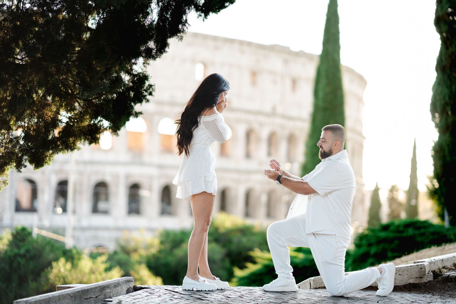 Man proposes to woman on one knee near the Colosseum in Rome, both dressed in white on a sunny day.