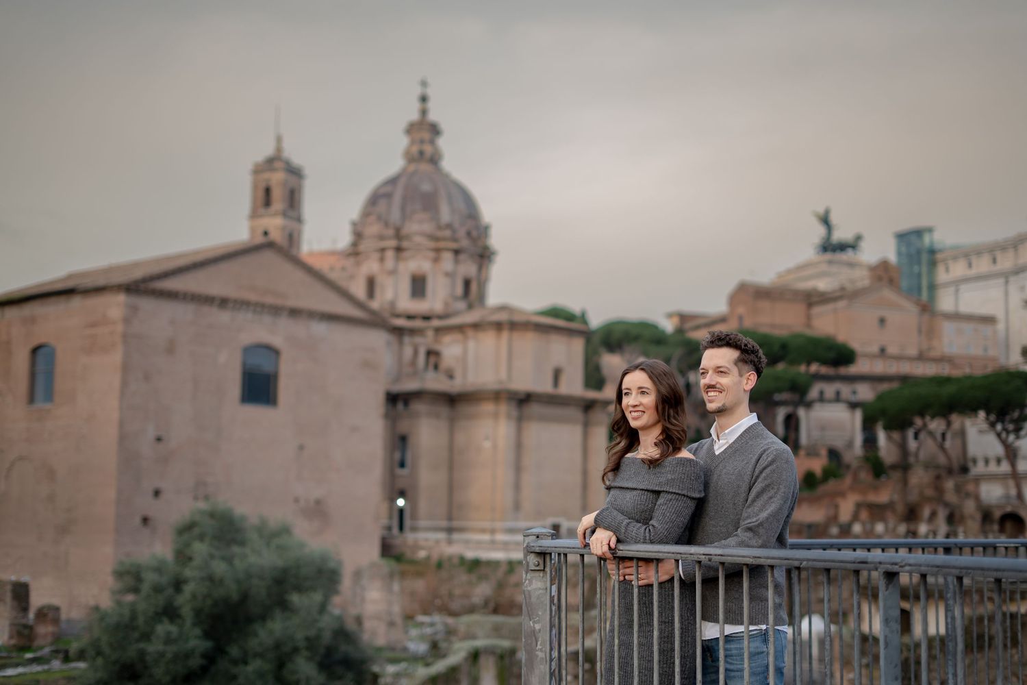 Couple posing at a railing with ancient Roman ruins and domed church in Rome, Italy backdrop.