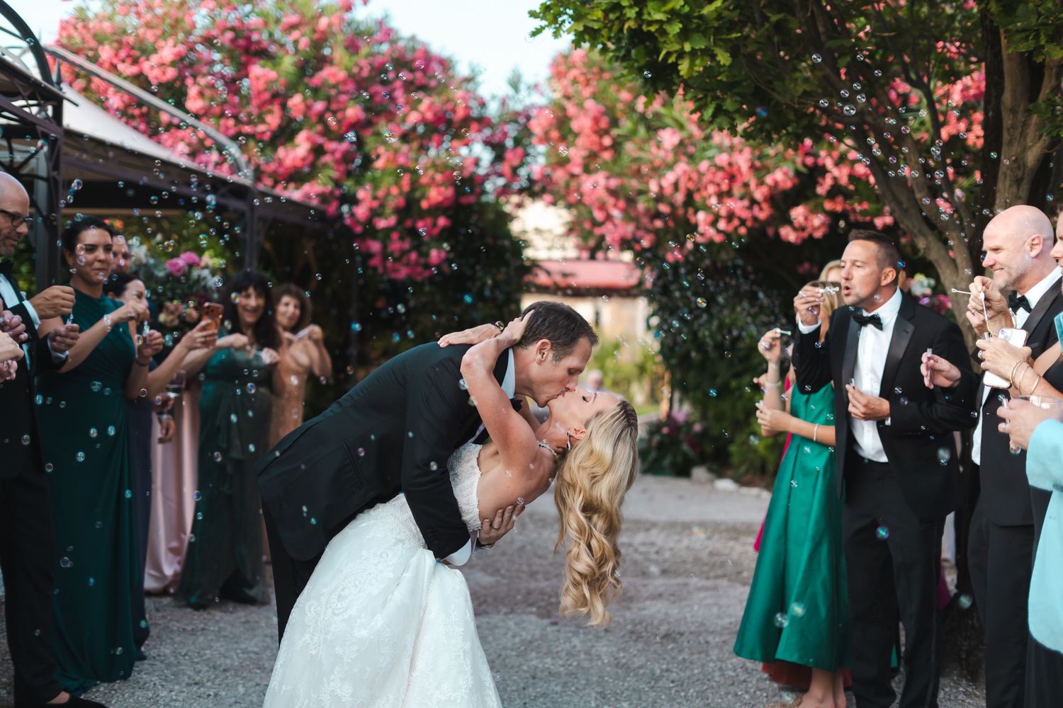 Groom dips bride for a kiss during outdoor wedding reception as guests celebrate with bubbles around them.
