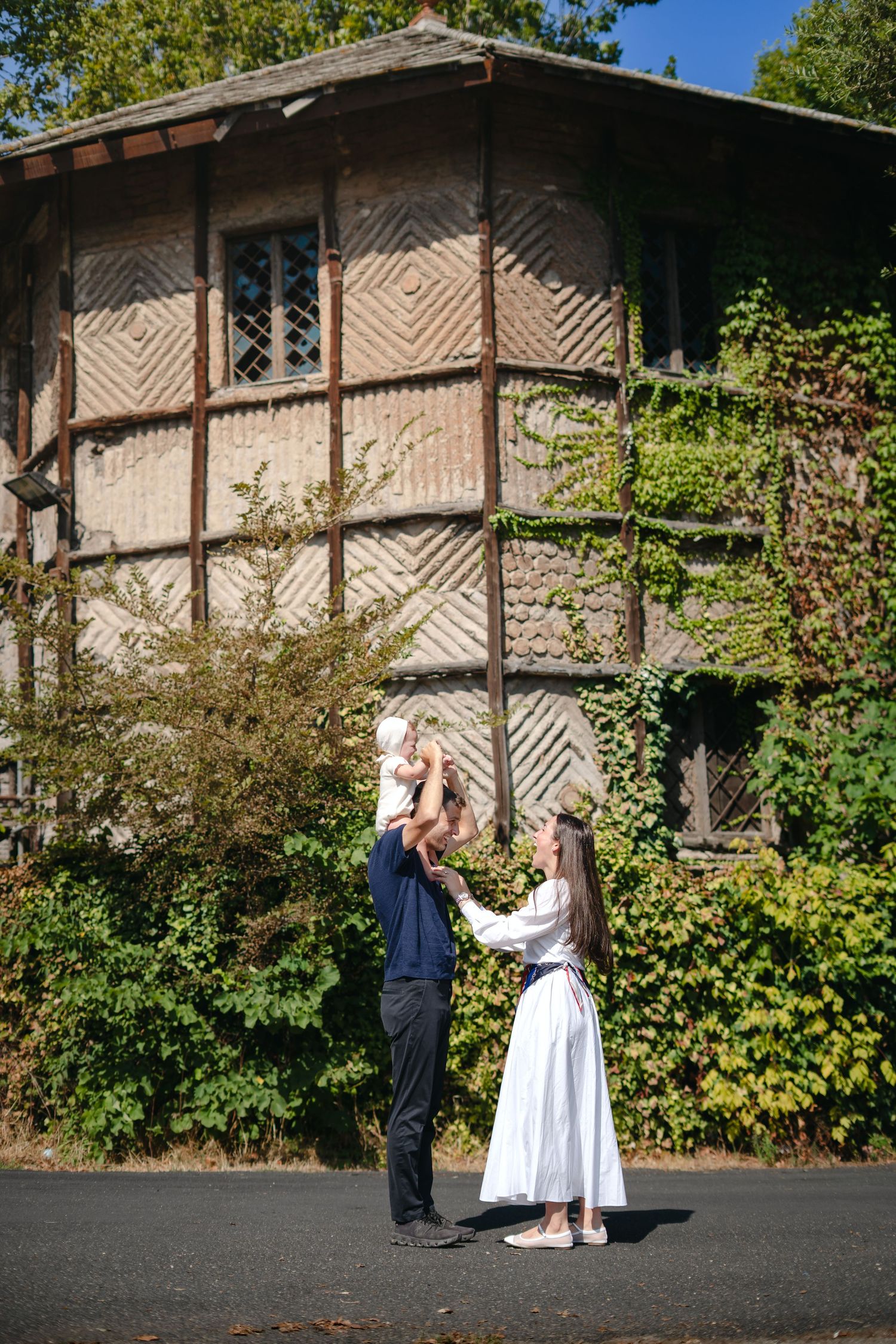 A happy family with a baby poses outdoors in front of a charming ivy-covered historic building on a sunny day.
