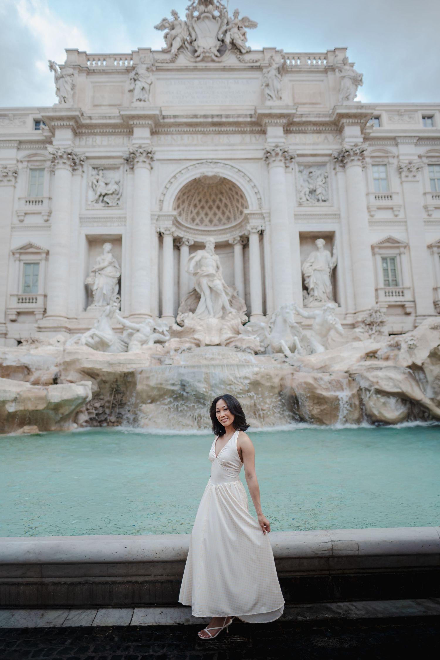 Woman in white dress posing at the Trevi Fountain in Rome, Italy, with baroque sculptures and turquoise water.