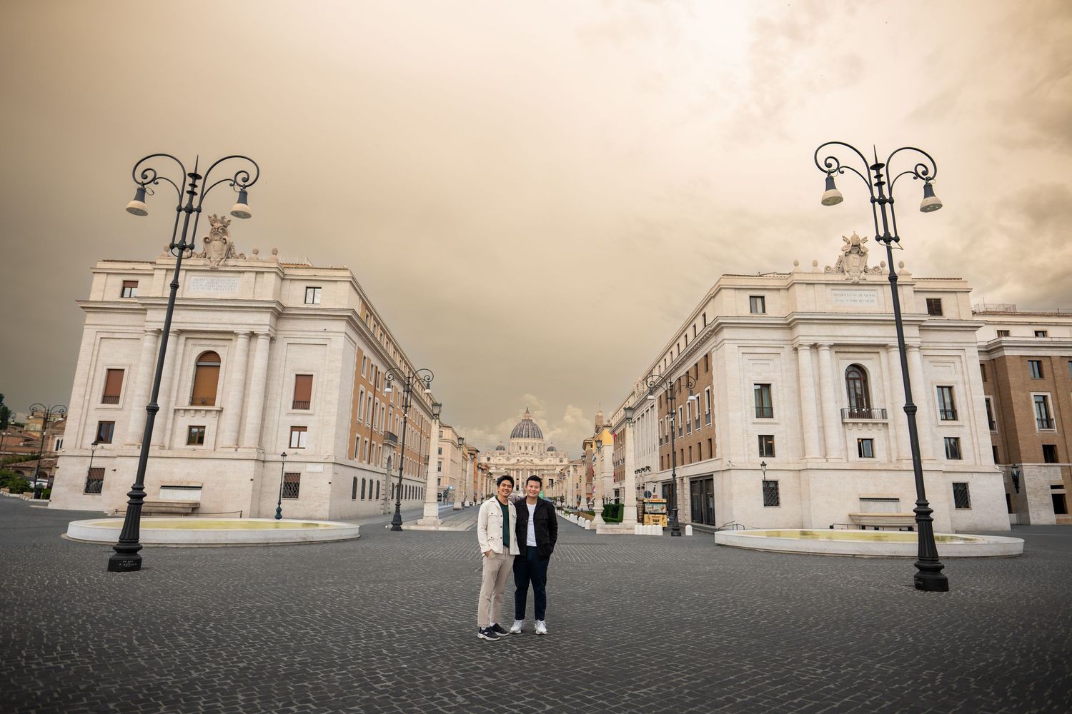 Two people pose on a grand cobblestone plaza in Rome, flanked by classical white buildings under a dramatic sky.