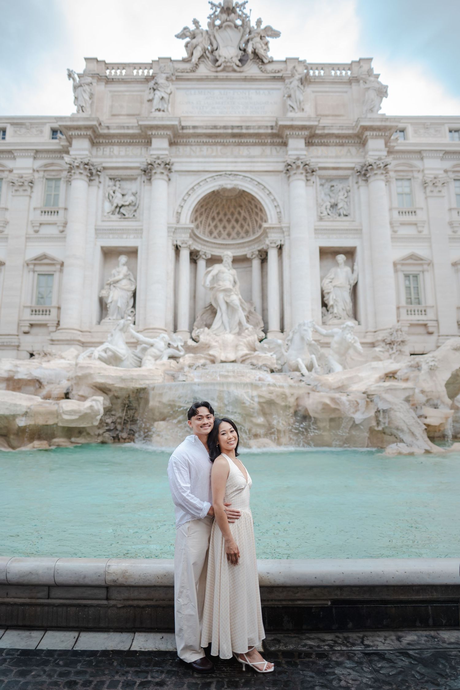 Couple in white attire posing romantically in front of the Trevi Fountain in Rome, Italy.