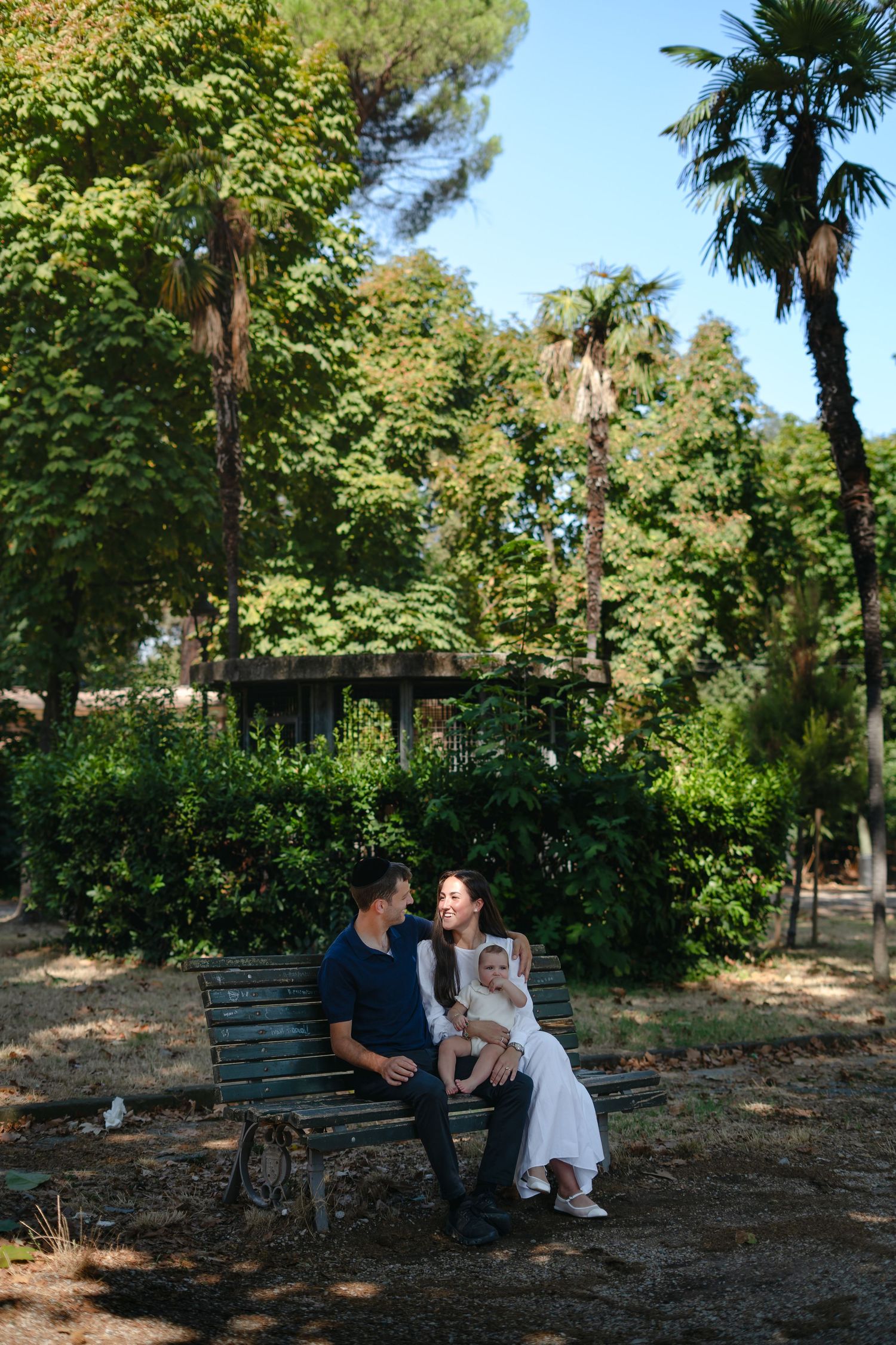 Family of three sitting on a park bench surrounded by lush green trees and palm trees on a sunny day.