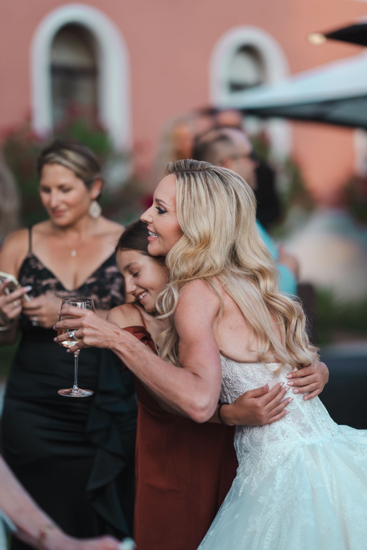 Blonde bride in lace dress embracing guest while holding wine glass at outdoor wedding reception.