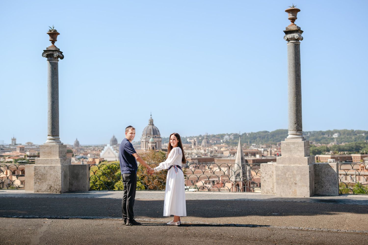 Couple holding hands on a rooftop terrace in Rome, Italy, with historic skyline and columns in the background.