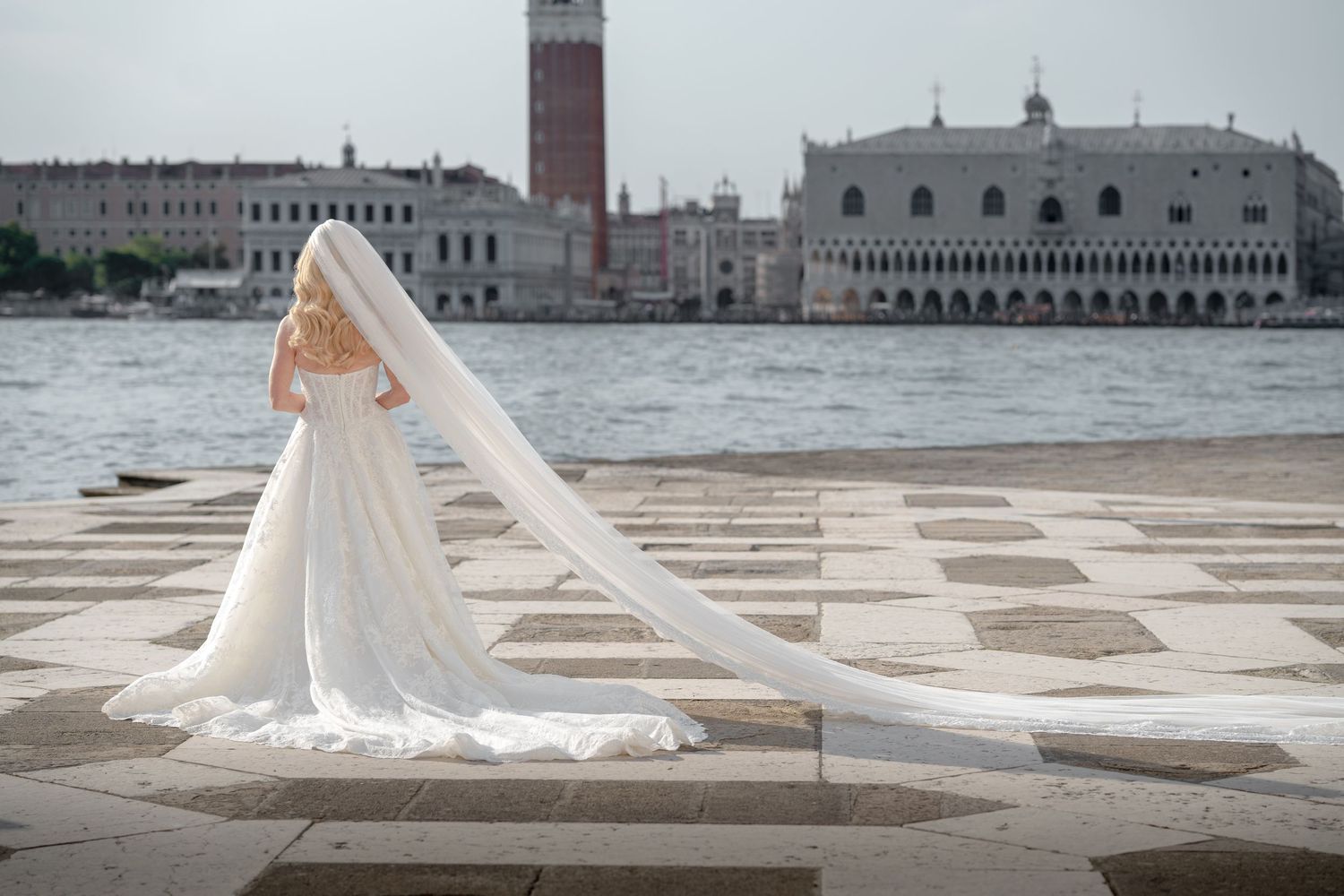 Bride in stunning white gown with long veil standing by Venice waterfront with St. Mark's Basilica backdrop.