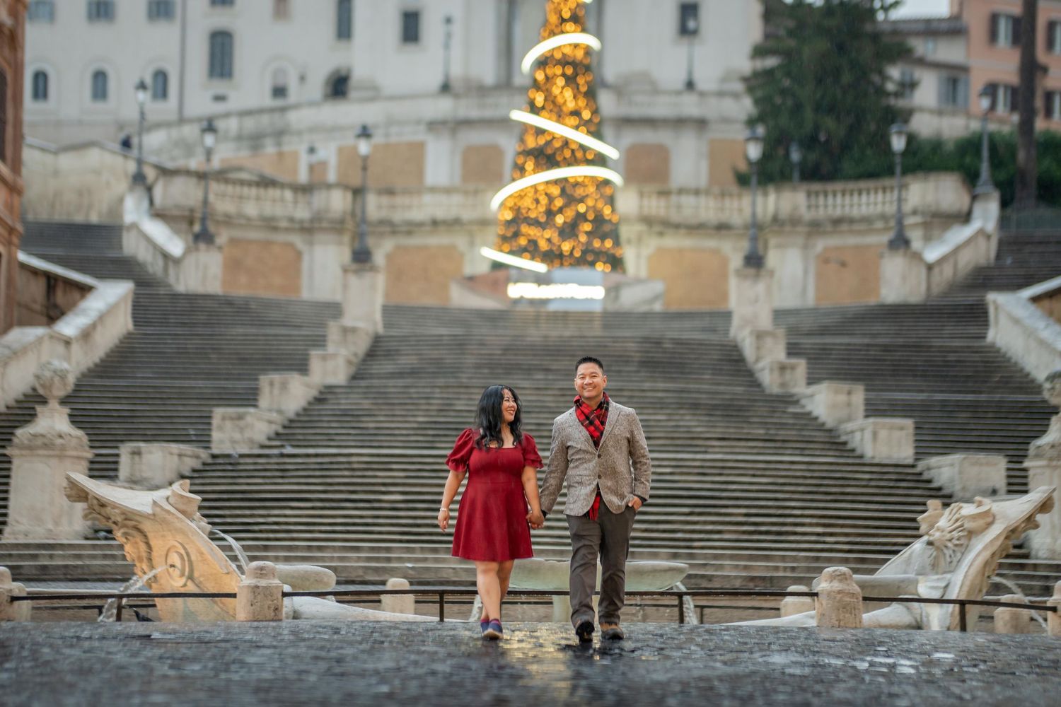 Couple holding hands at the Spanish Steps in Rome with a glowing Christmas tree backdrop during the holiday season.