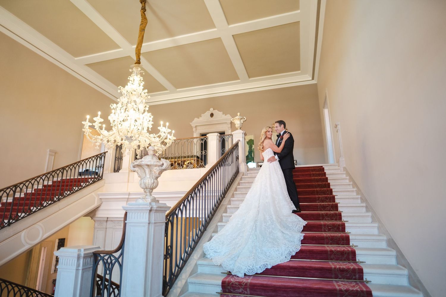 Elegant bride and groom posing on grand staircase with chandelier in a luxurious wedding venue.