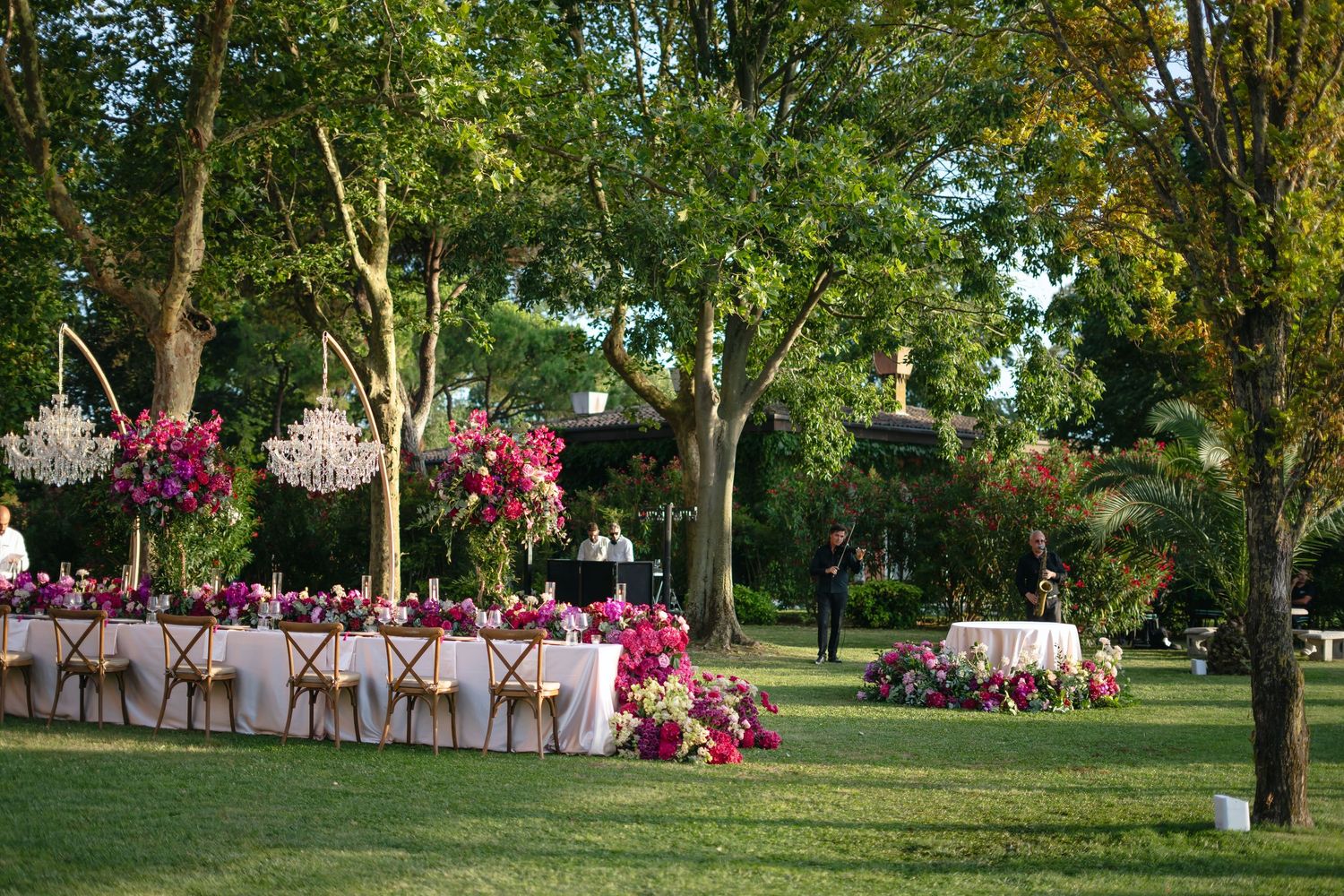 Elegant outdoor wedding reception with long tables, crystal chandeliers, and vibrant pink floral arrangements in a garden.