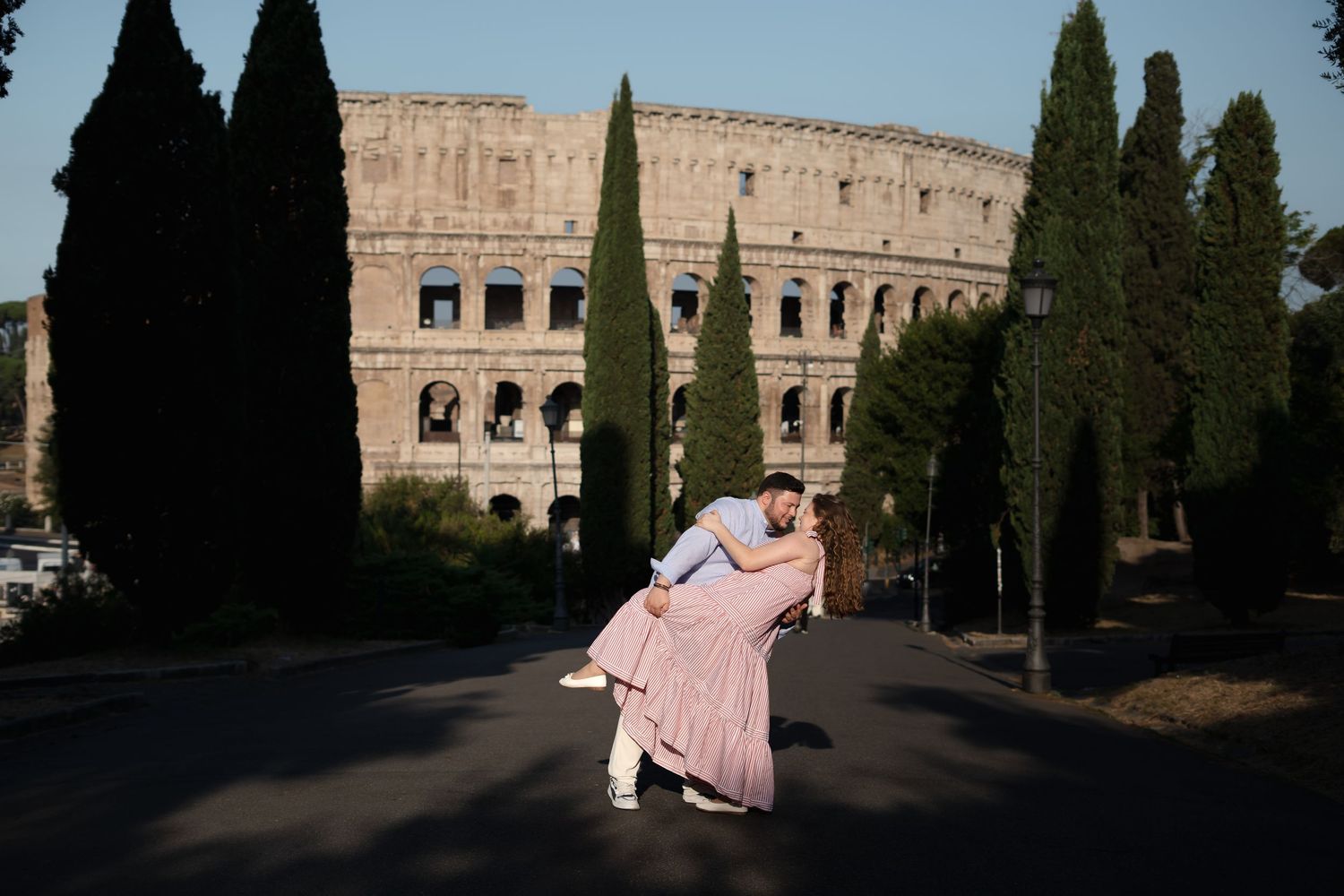 Couple dancing romantically in front of the Colosseum in Rome, Italy, surrounded by tall cypress trees.