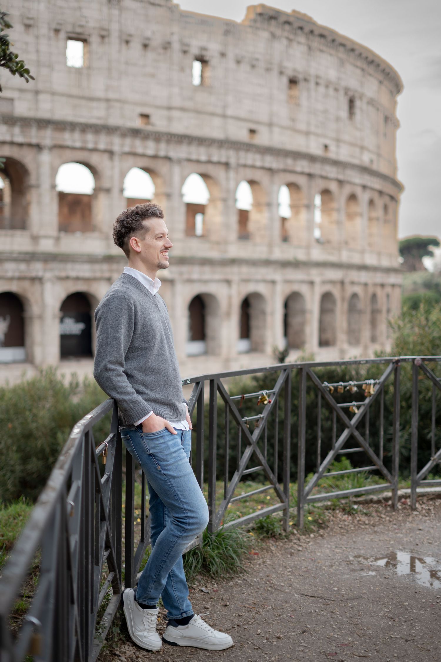 Man in gray sweater and jeans smiling while leaning on a fence with the Roman Colosseum in the background.