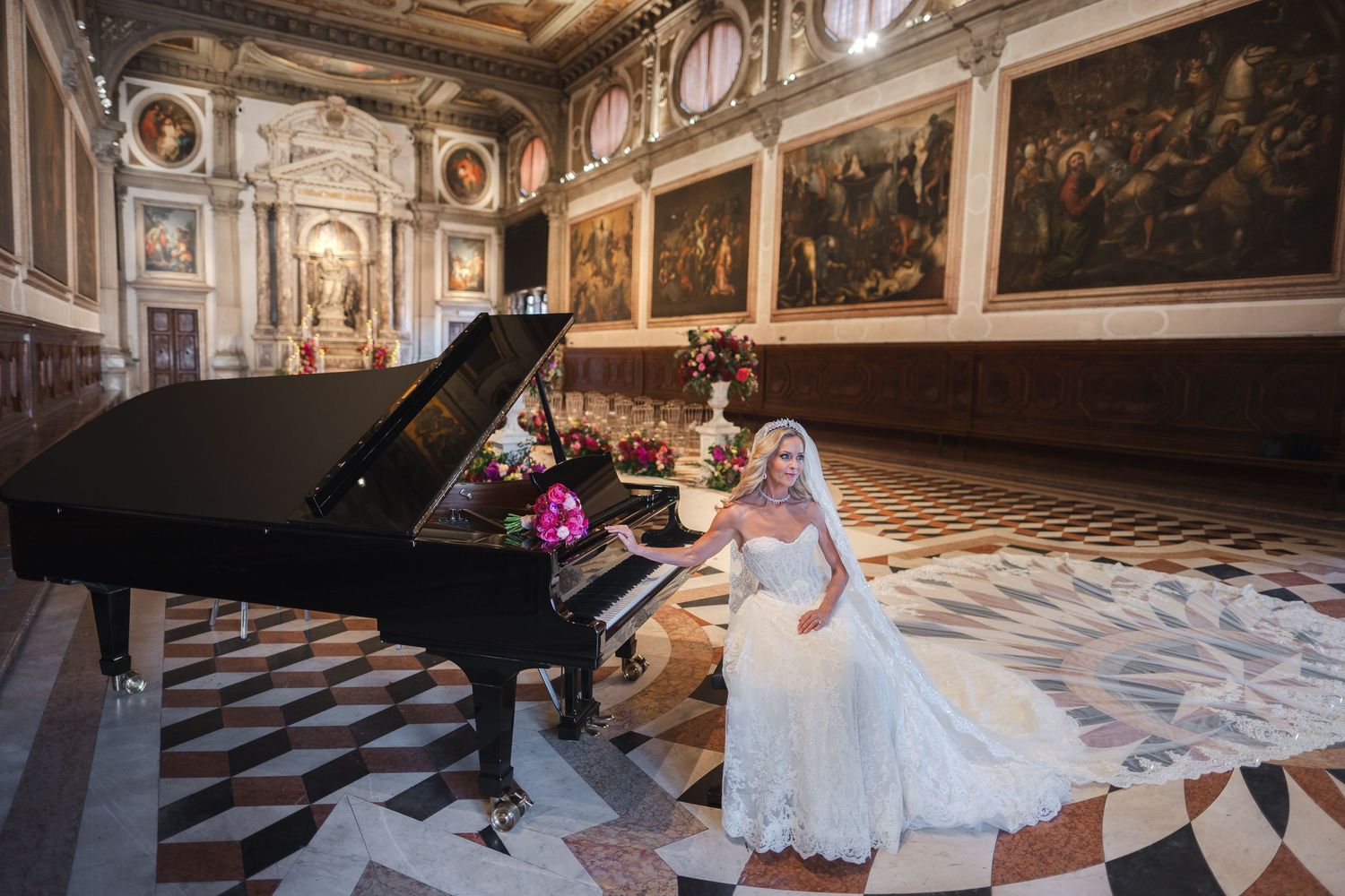 Bride in white gown seated by a grand piano in an ornate hall with classical paintings and marble floors.