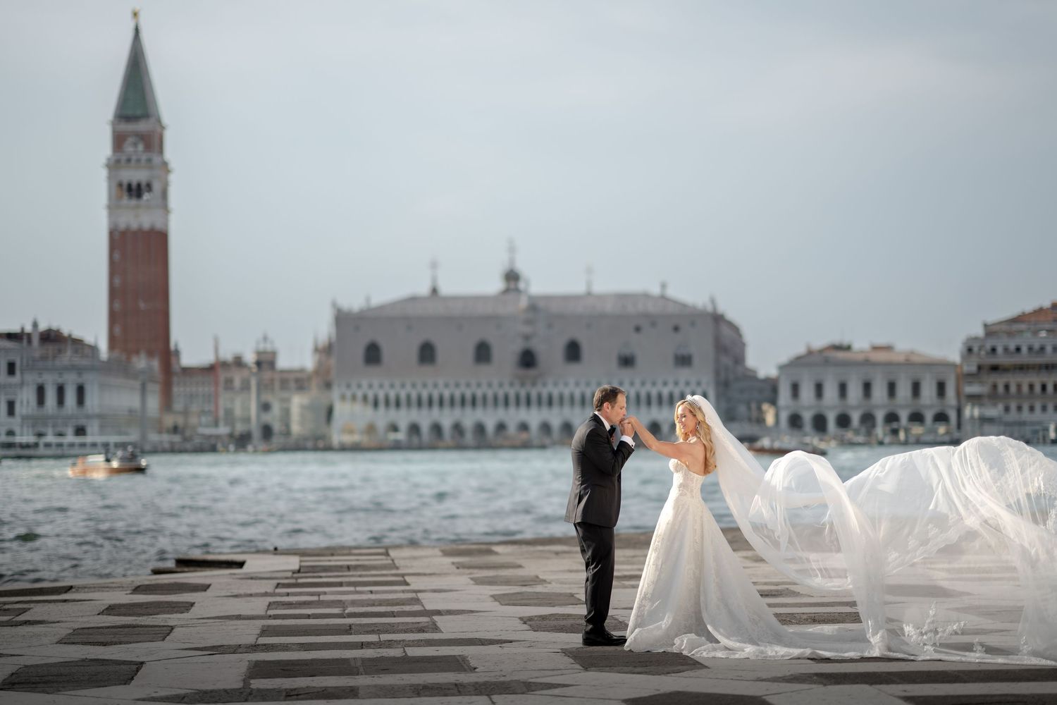 Wedding couple embracing on Venice lagoon pier with flowing veil and St. Mark's Square skyline.