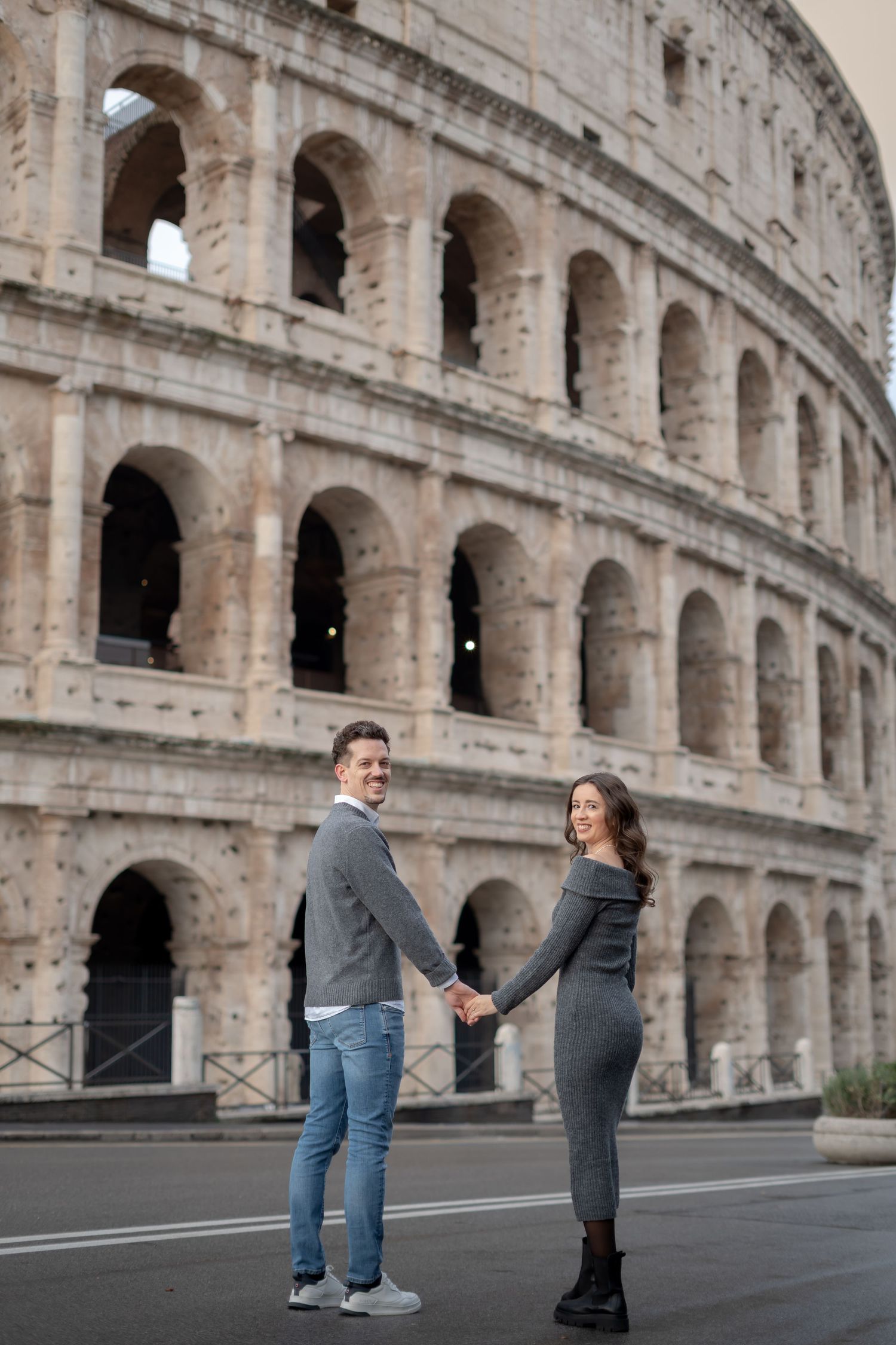 Couple holding hands in front of the Colosseum in Rome, Italy, wearing matching gray outfits on a sunny day.