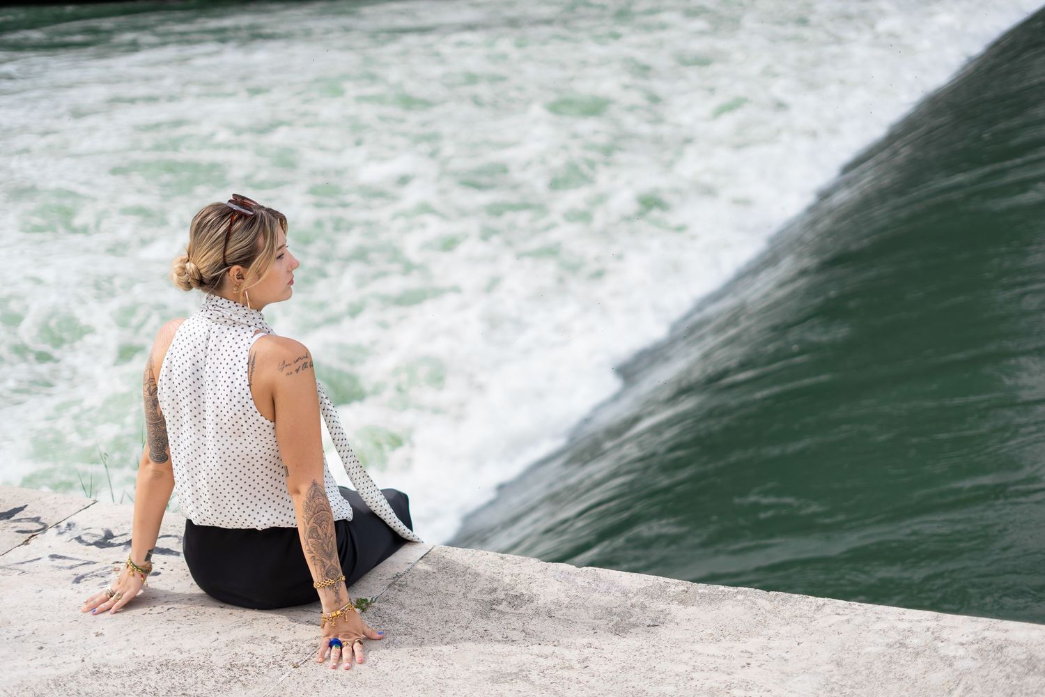 Woman with tattoos sitting on stone ledge beside rushing water, wearing polka dot top and black pants.