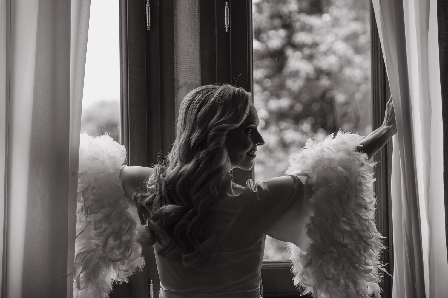 Black and white photo of bride in feathered robe gazing out an open window before her wedding.