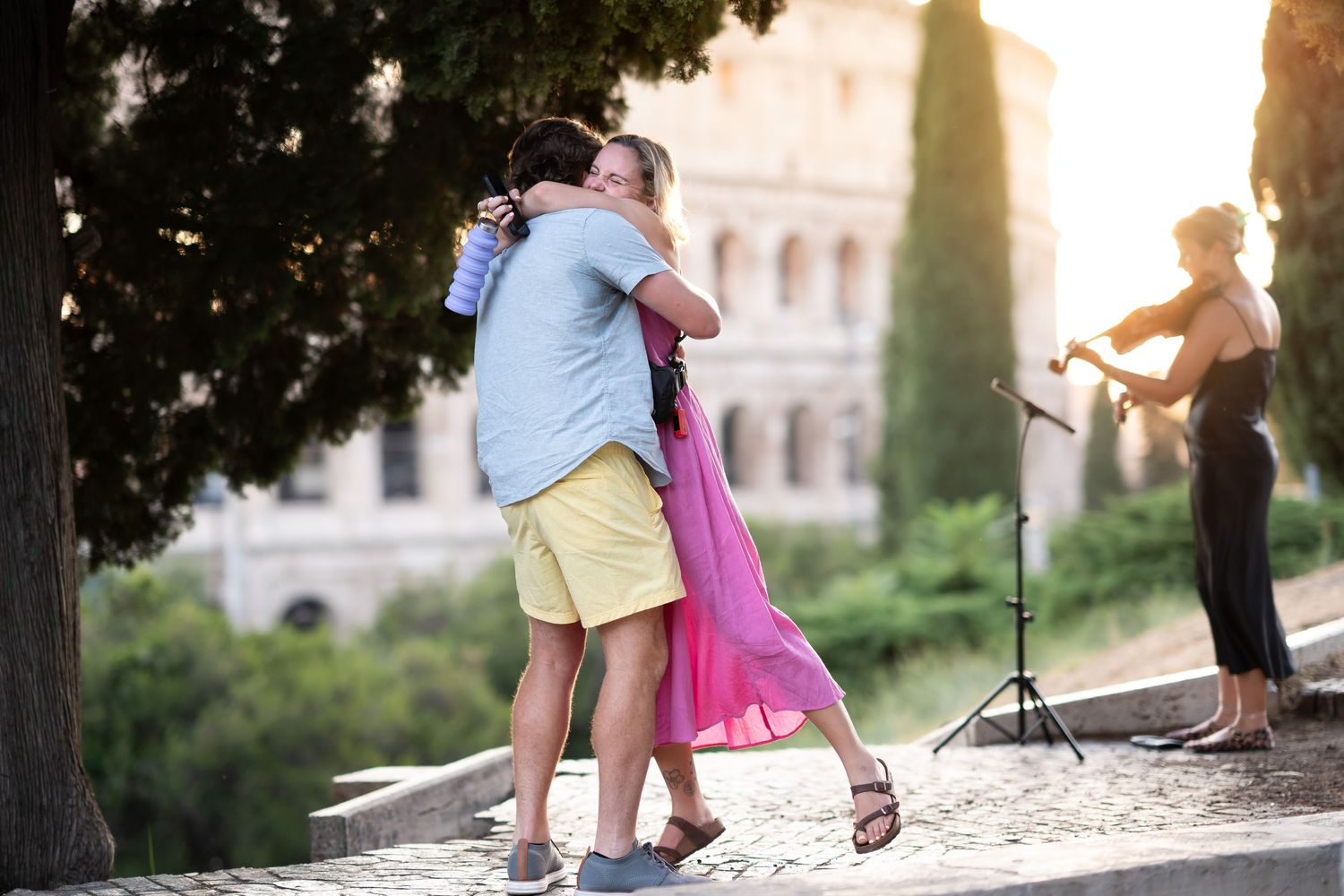 Couple embracing near the Colosseum in Rome with a violinist performing in the background at sunset.