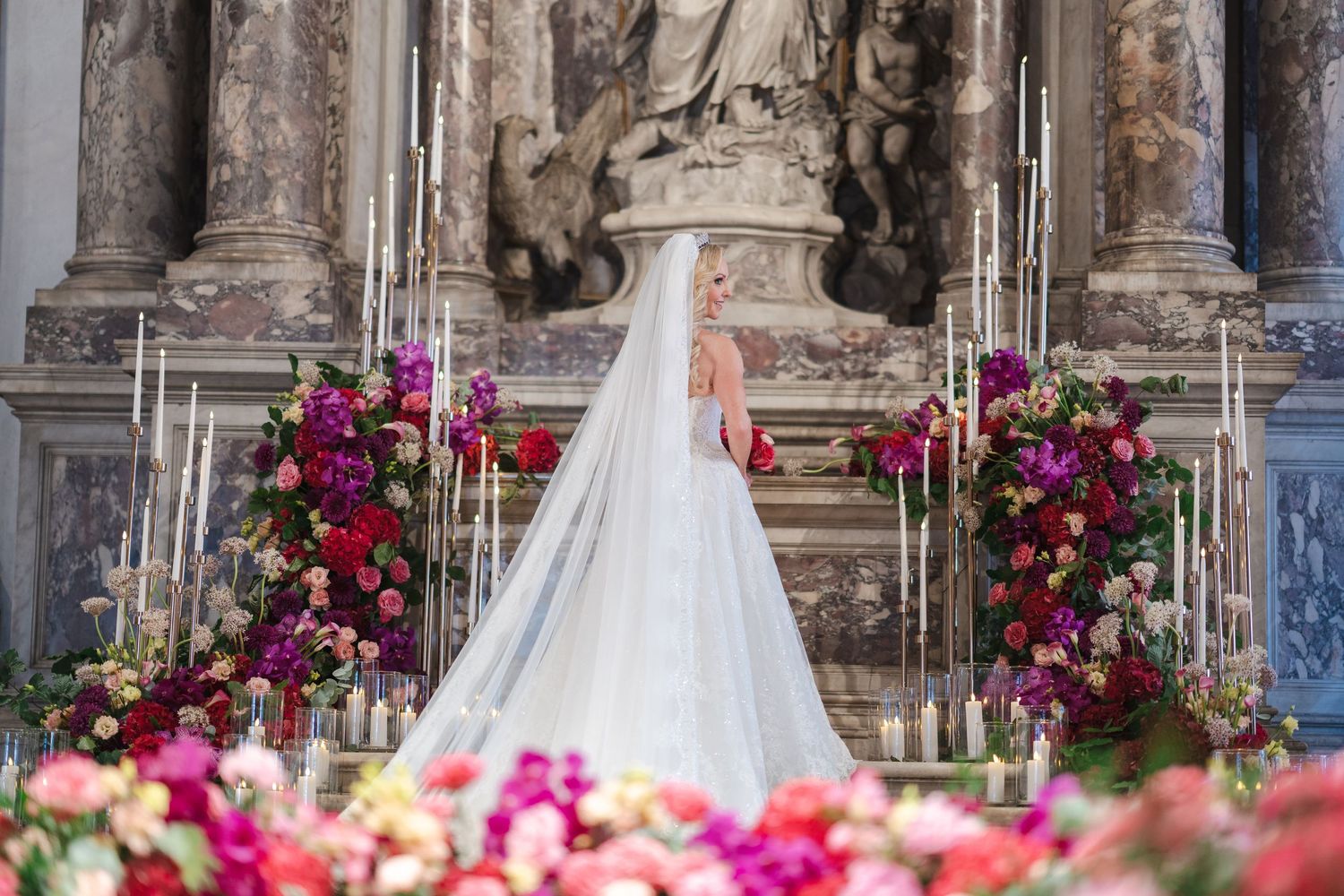 Bride in stunning gown and cathedral veil stands before elaborate floral altar in Venice church.