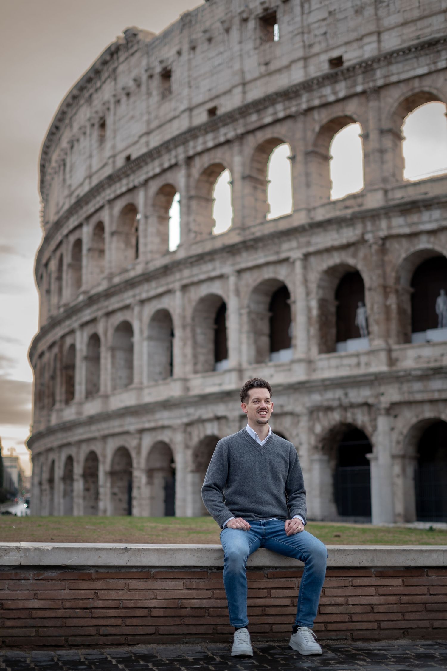 Smiling man sitting on a wall in front of the iconic Colosseum in Rome, Italy.