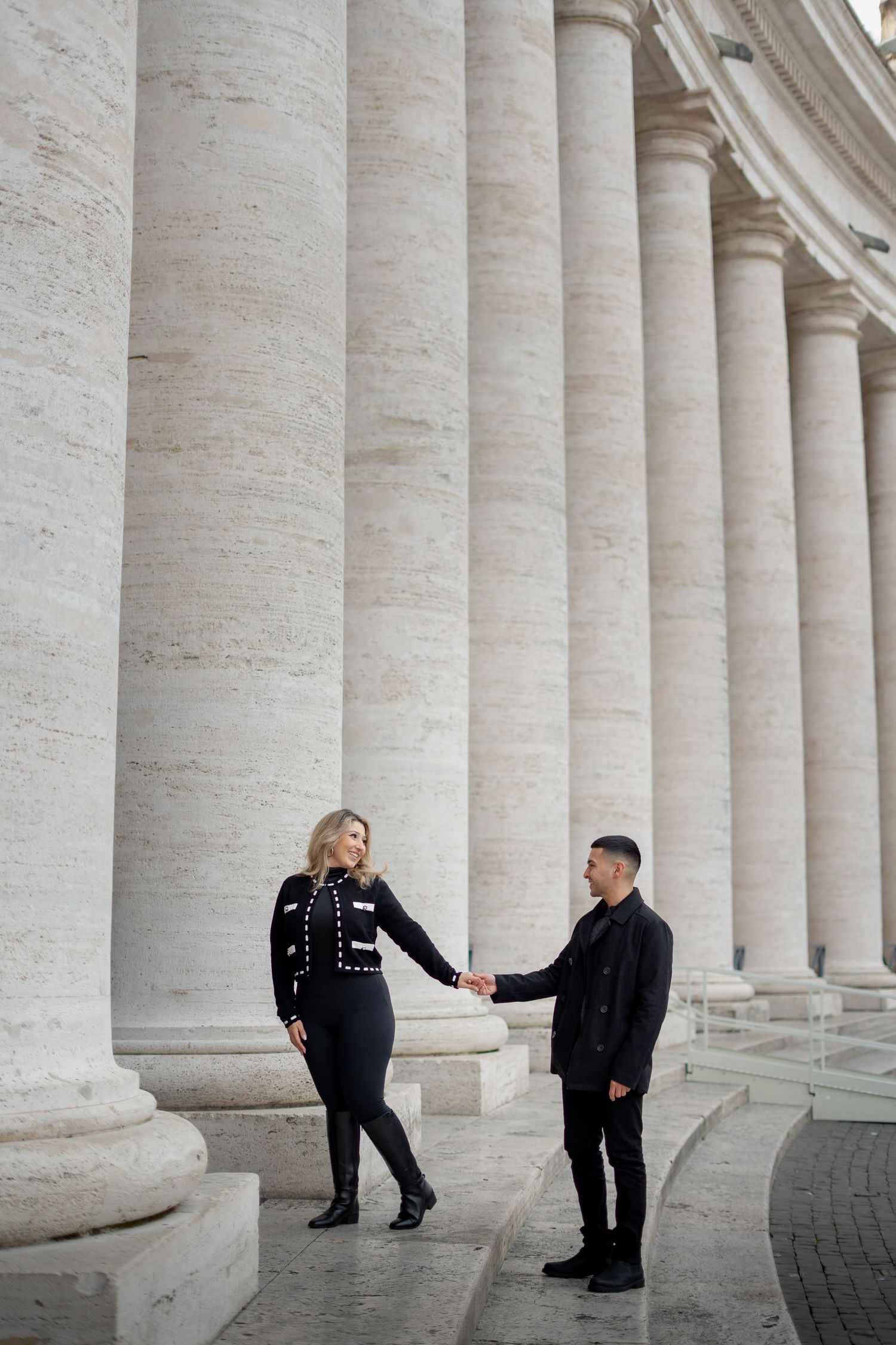 Couple in black outfits holding hands among tall marble columns at St. Peters Square Vatican Rome.