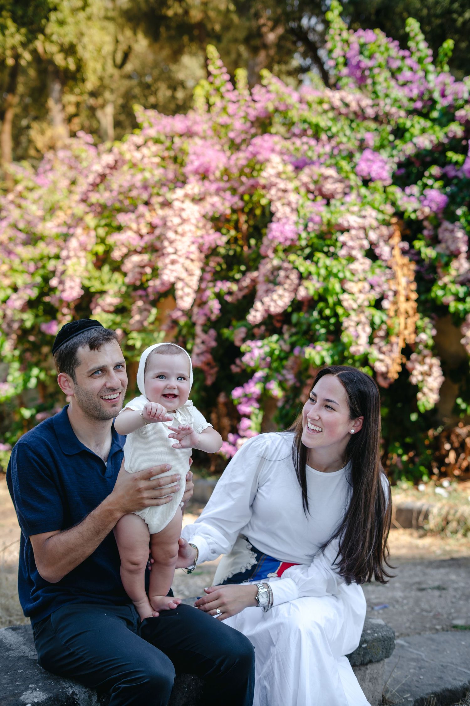 Happy family with baby sitting outdoors among pink flowering bushes, parents smiling at infant in white outfit.