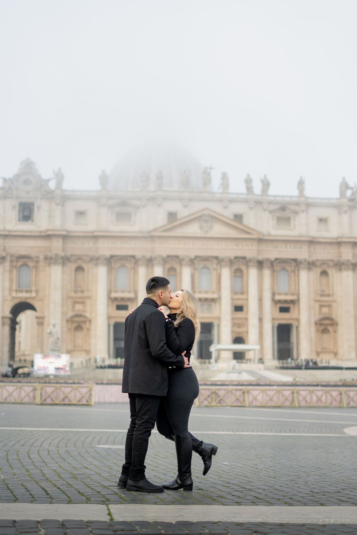 Couple kissing in St. Peter's Square, Vatican City, on a foggy day, both dressed in elegant black outfits.