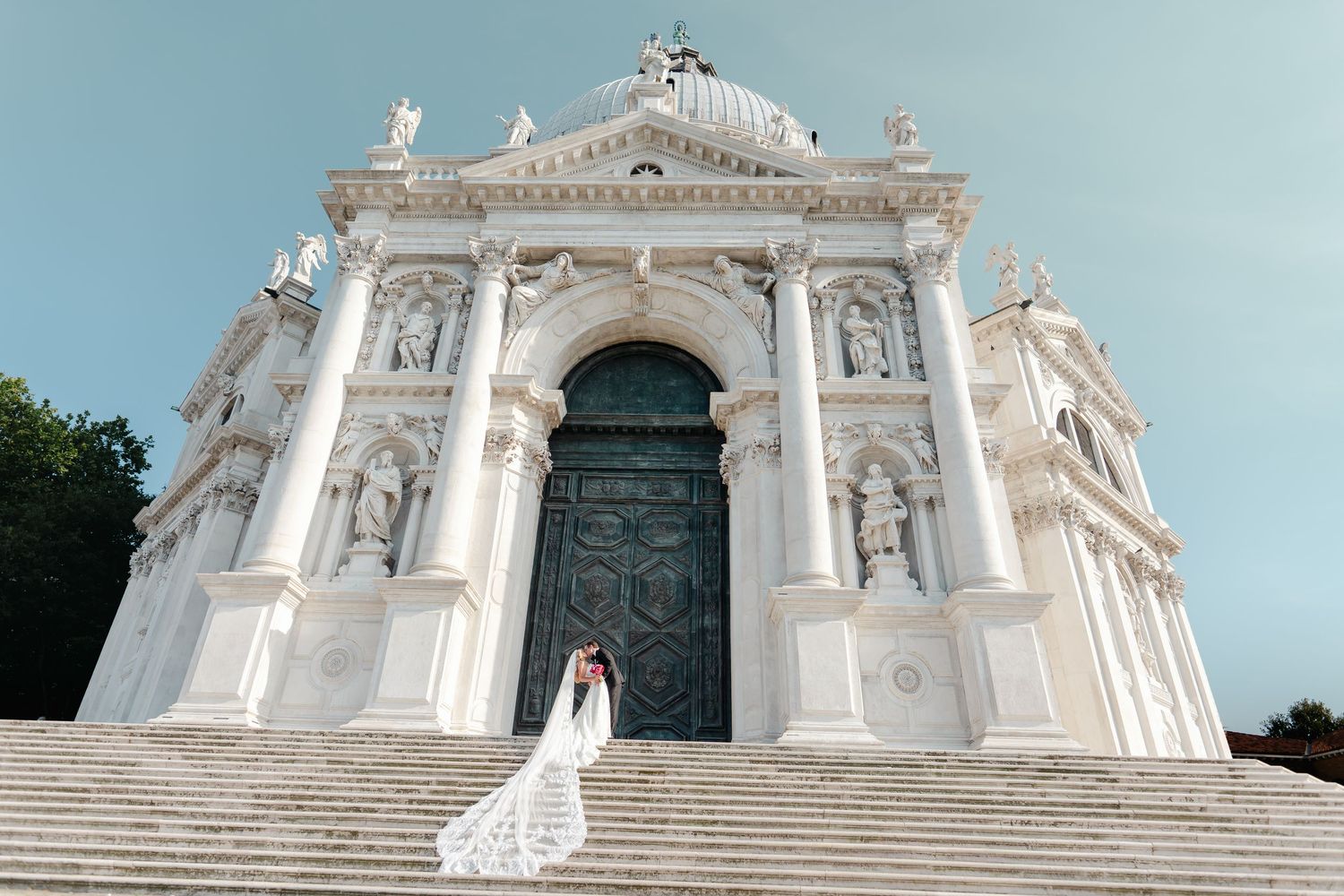 Bride and groom embrace on the steps of a grand white baroque church in Venice, Italy.
