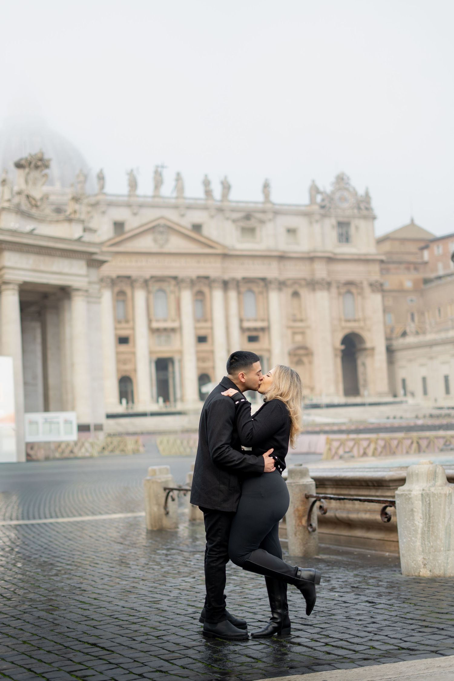 Couple sharing a romantic kiss in St. Peter's Square, Vatican City, dressed in elegant black outfits on a misty day.