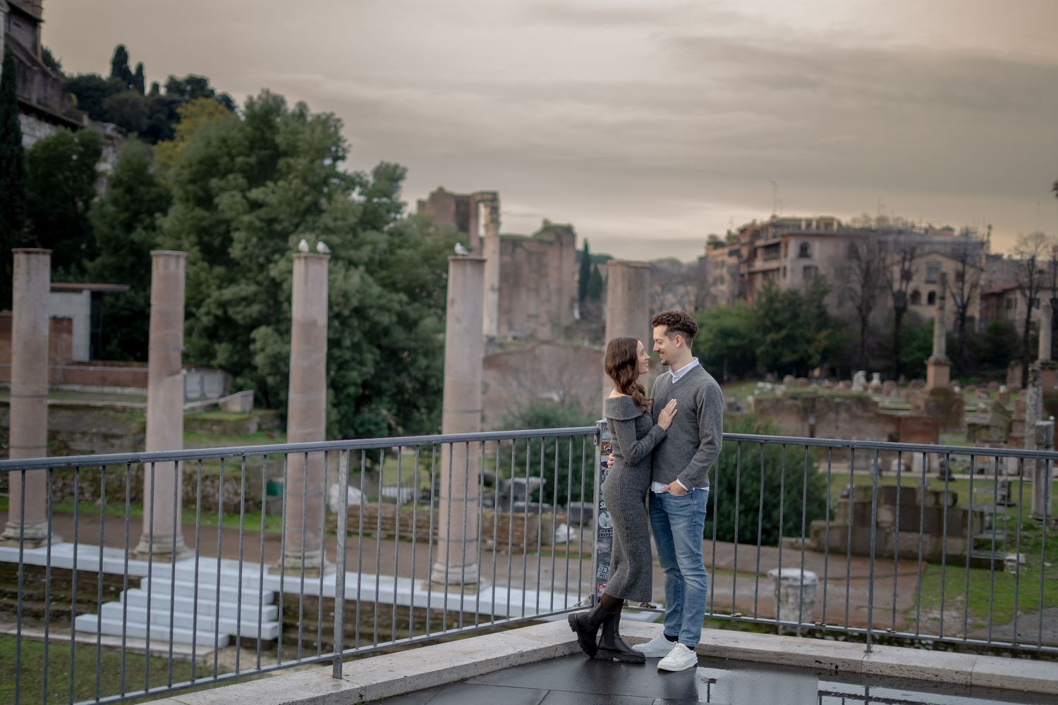 Couple embracing near ancient Roman ruins on a cloudy day, with historic columns and architecture in the background.