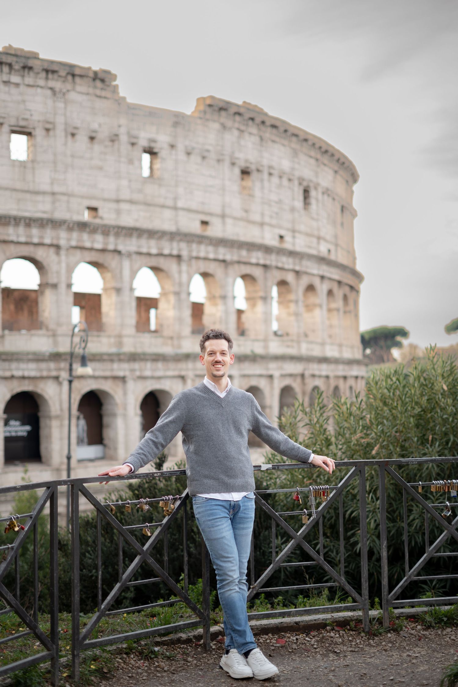 Man smiling in front of the Colosseum in Rome, Italy, wearing a gray sweater and jeans on a cloudy day.