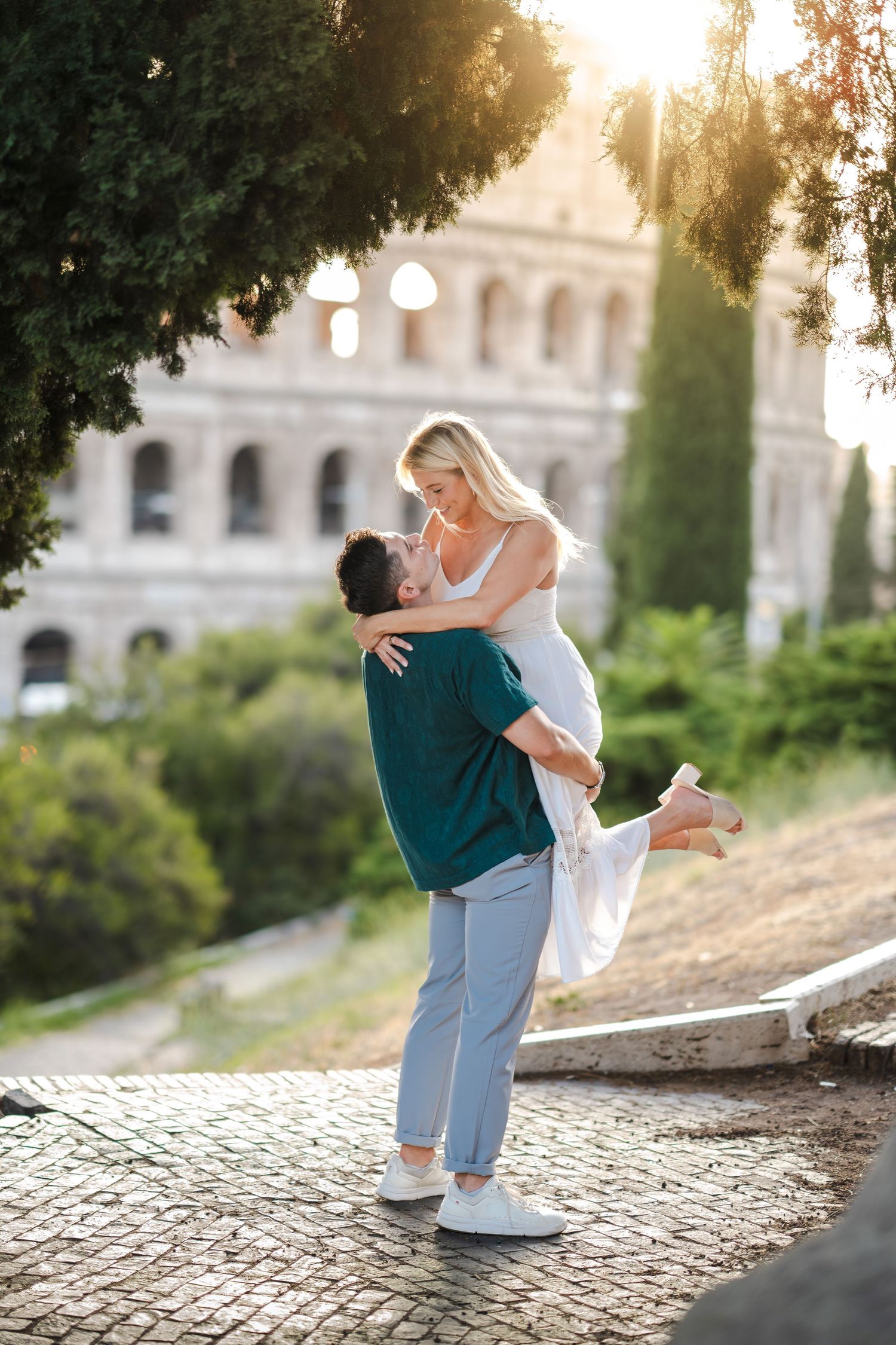 Couple embracing in front of the Colosseum in Rome at golden hour, man lifting woman in white dress.