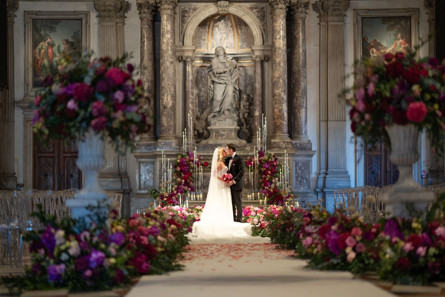 Bride and groom kiss at ornate church altar surrounded by lush pink and red floral arrangements.