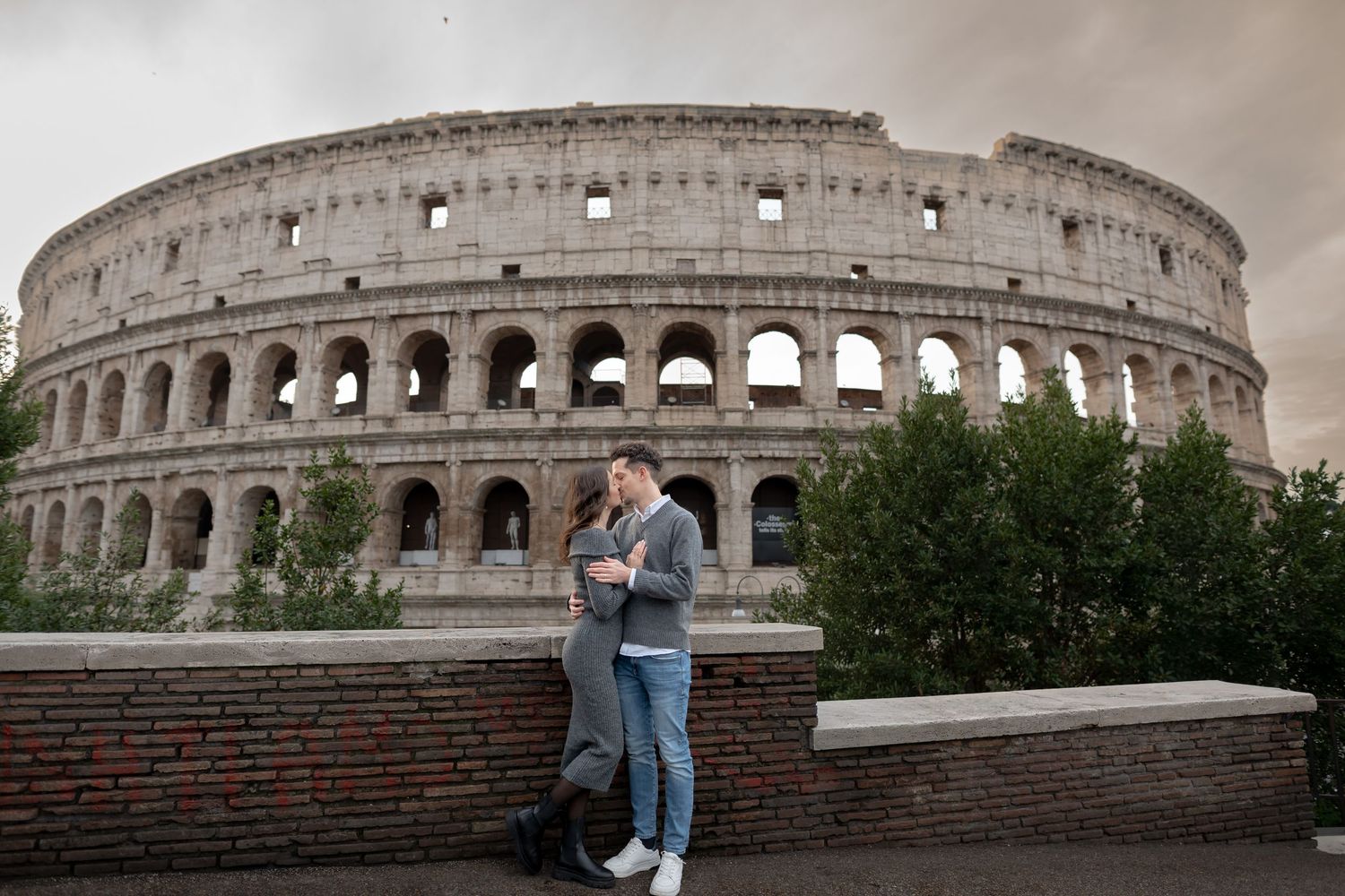Romantic couple embracing in front of the ancient Colosseum in Rome under a dramatic cloudy sky.