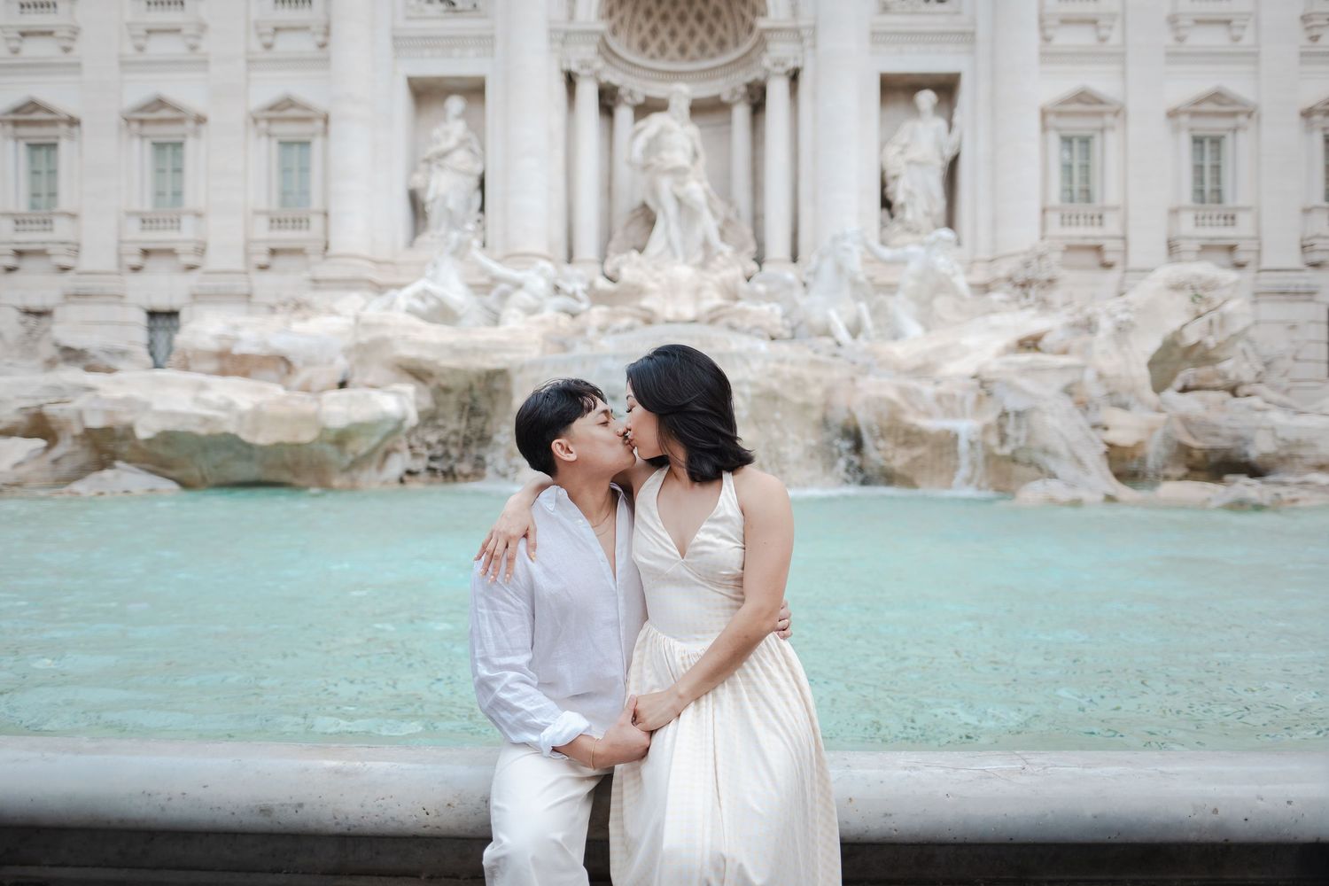 Couple sharing a romantic kiss in front of the Trevi Fountain in Rome, dressed in elegant white outfits.
