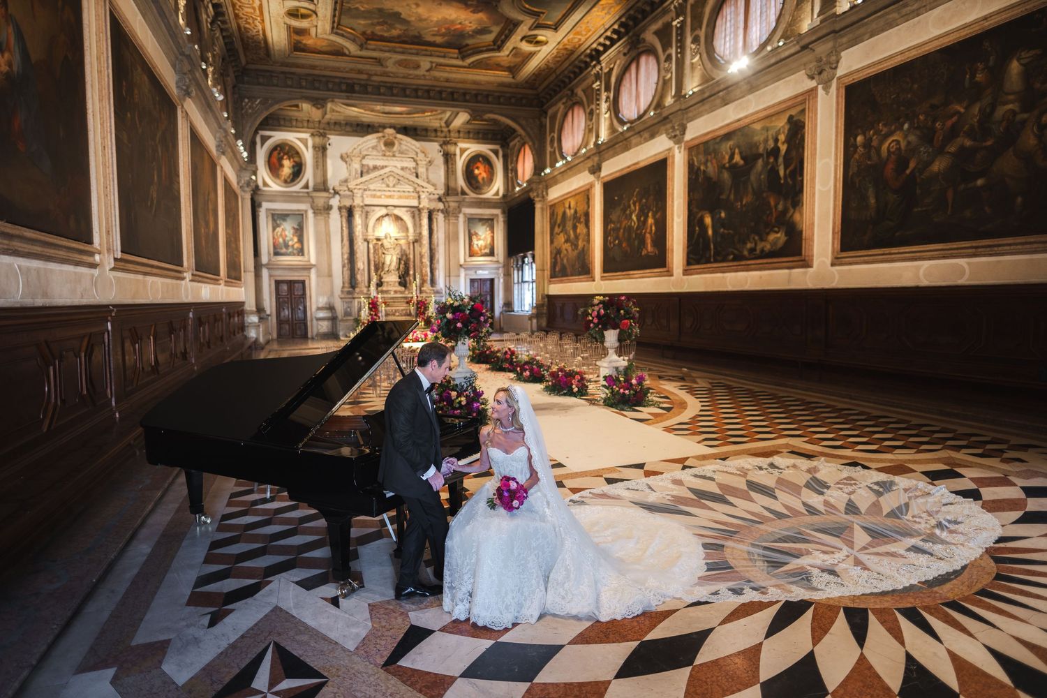 Bride and groom sit by a grand piano in an ornate Venetian hall with gilded paintings and marble floors.