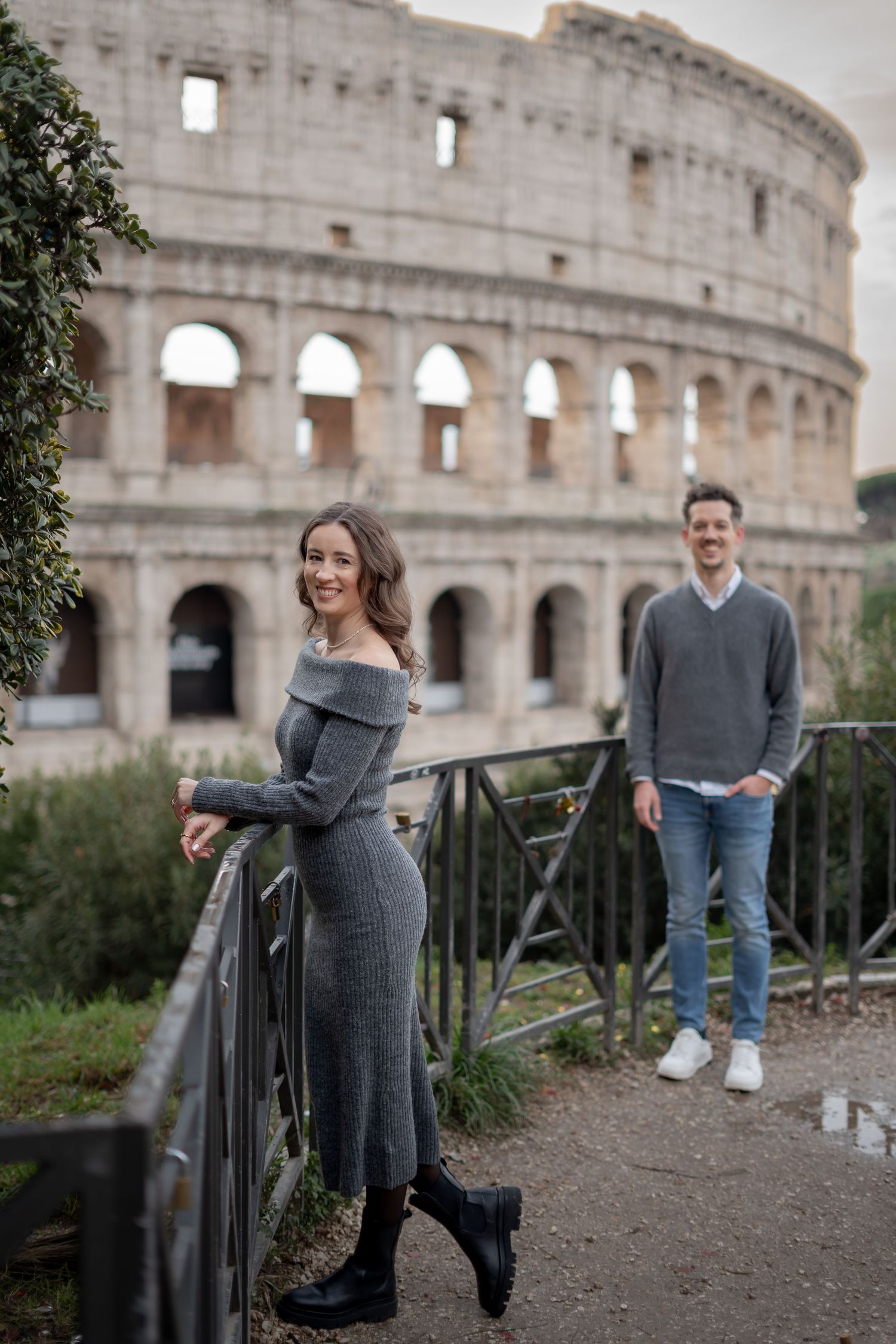Couple posing near the Colosseum in Rome, woman in gray dress smiling at camera, man in gray sweater standing behind.