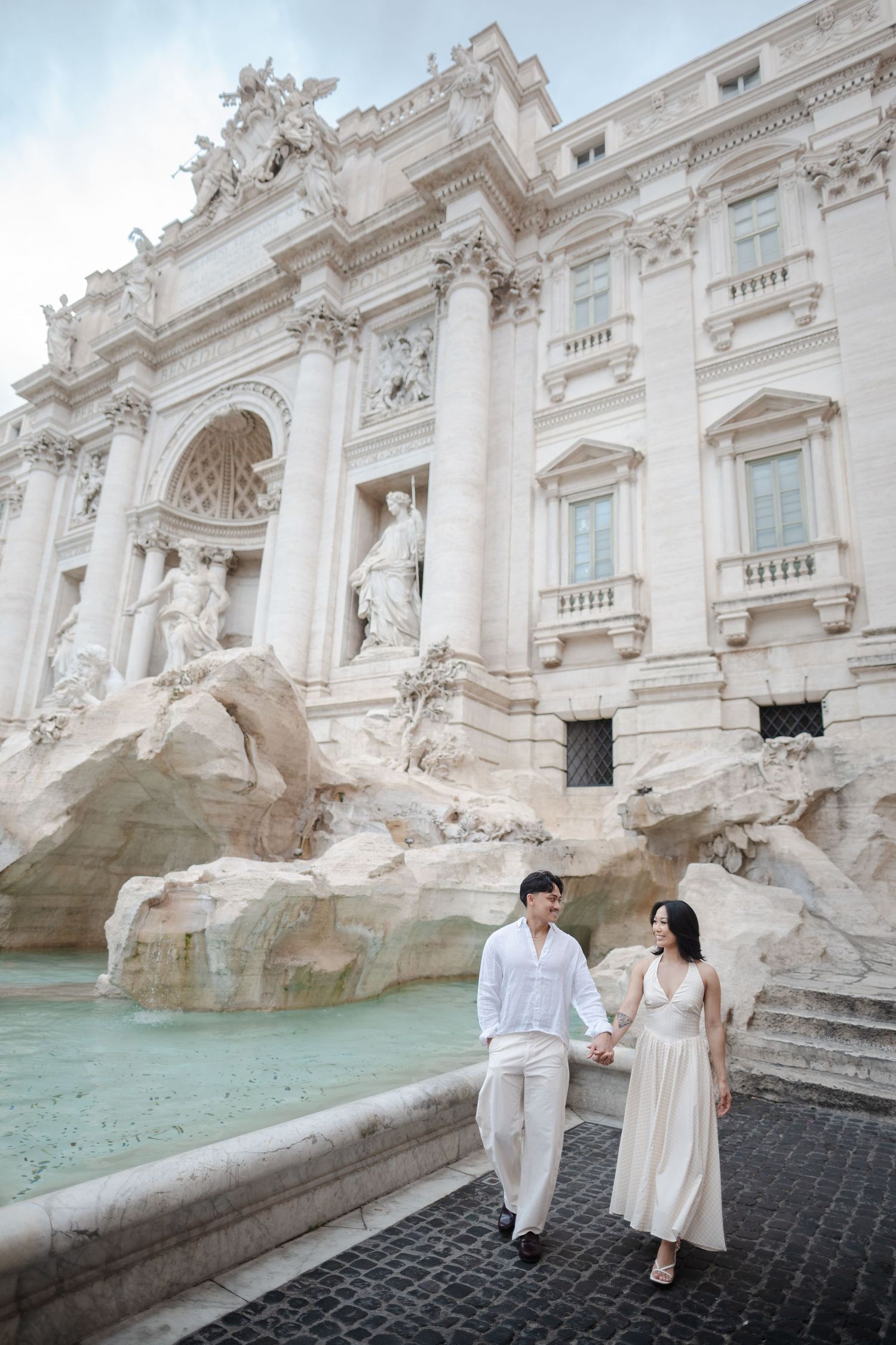Couple holding hands at the Trevi Fountain in Rome, Italy, with baroque architecture and sculptures in the background.