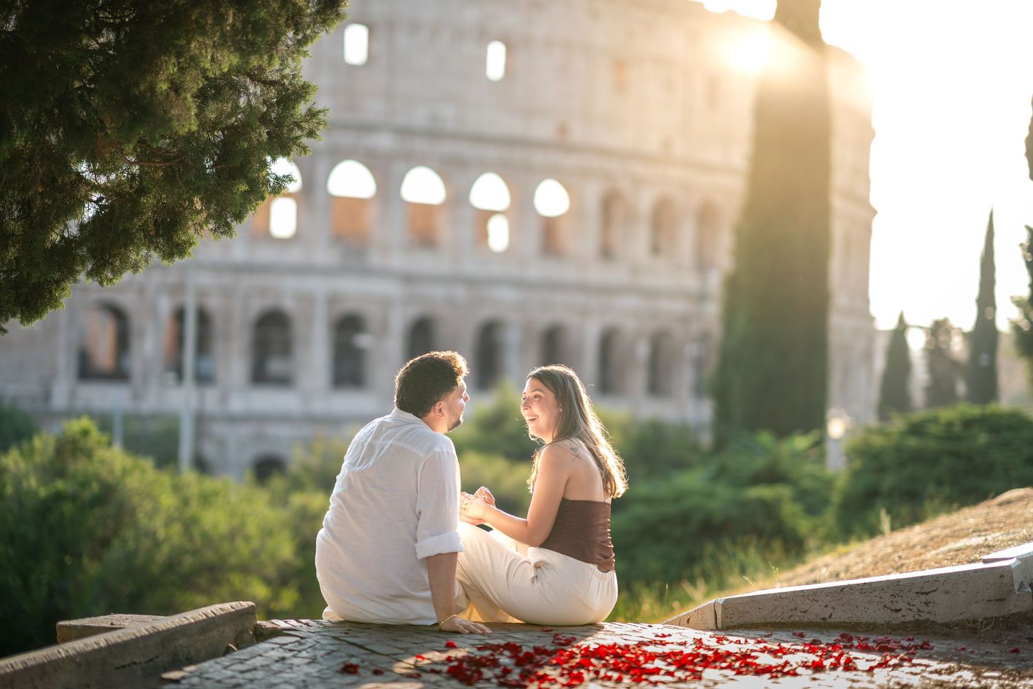Couple sitting on steps with red rose petals in front of the Colosseum in Rome at golden hour sunset.